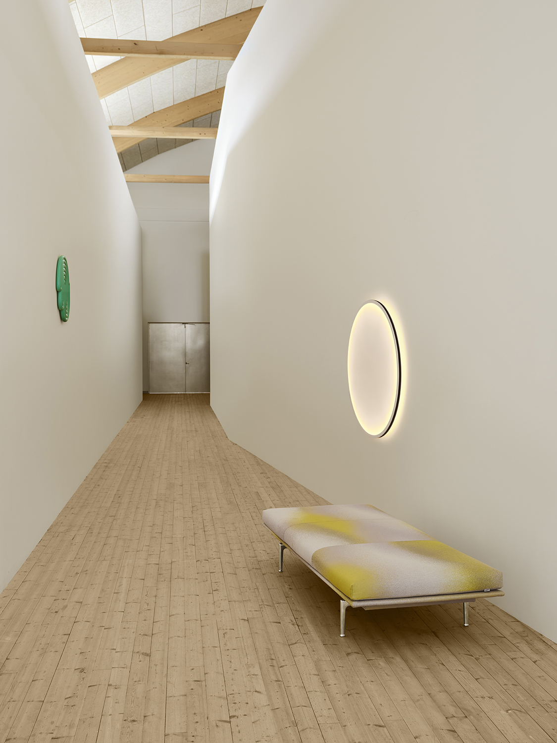 Outline Daybed is placed in a hall with light wooden floors and white walls. A round wall lamp hangs above, casting a soft halo of light that adds a calm atmosphere to the space.