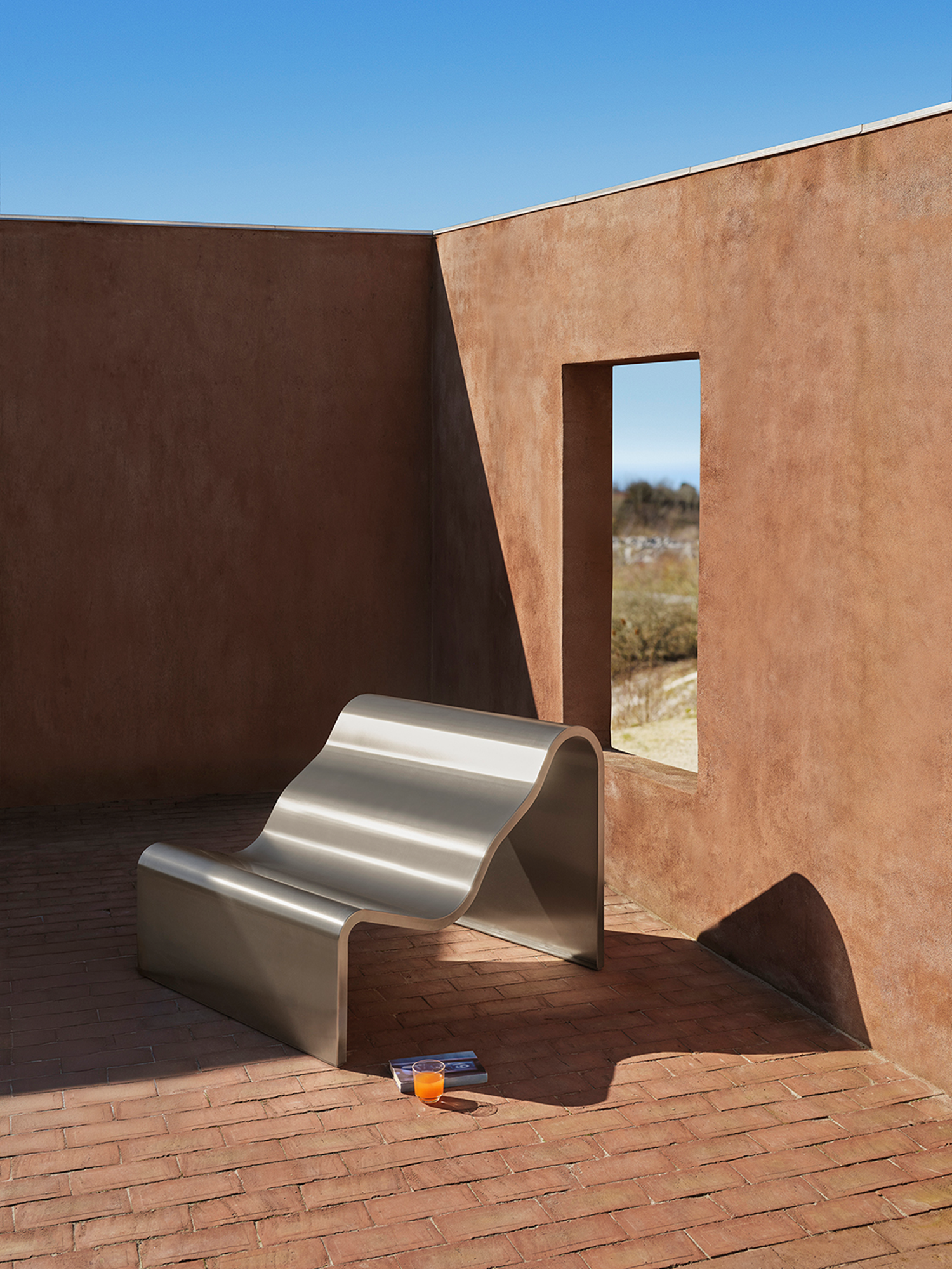 A architectural metal bench with a wavy, sculptural design is placed outdoor. A glass in a clear color is placed beside the bench. 