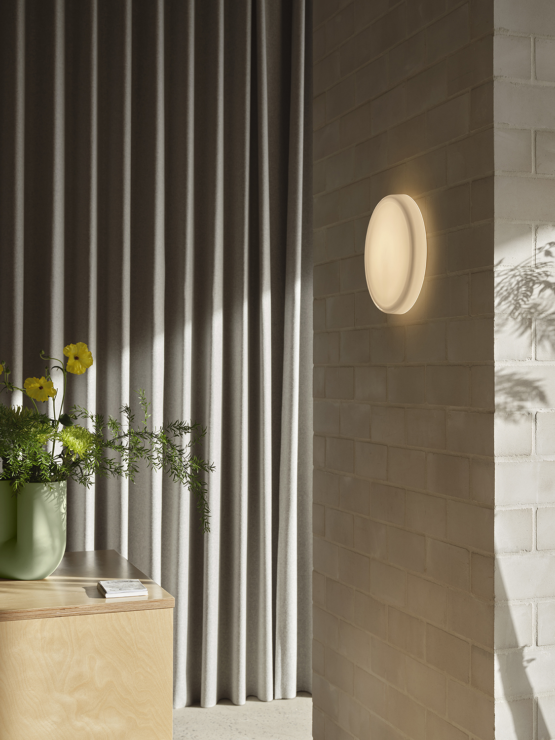 A minimalistich hallway featuring brushed stone walls. A round wall lamp hanging on the wall and a sculptural vase in light green is placed on a wooden table, creating a serene and modern atmosphere.