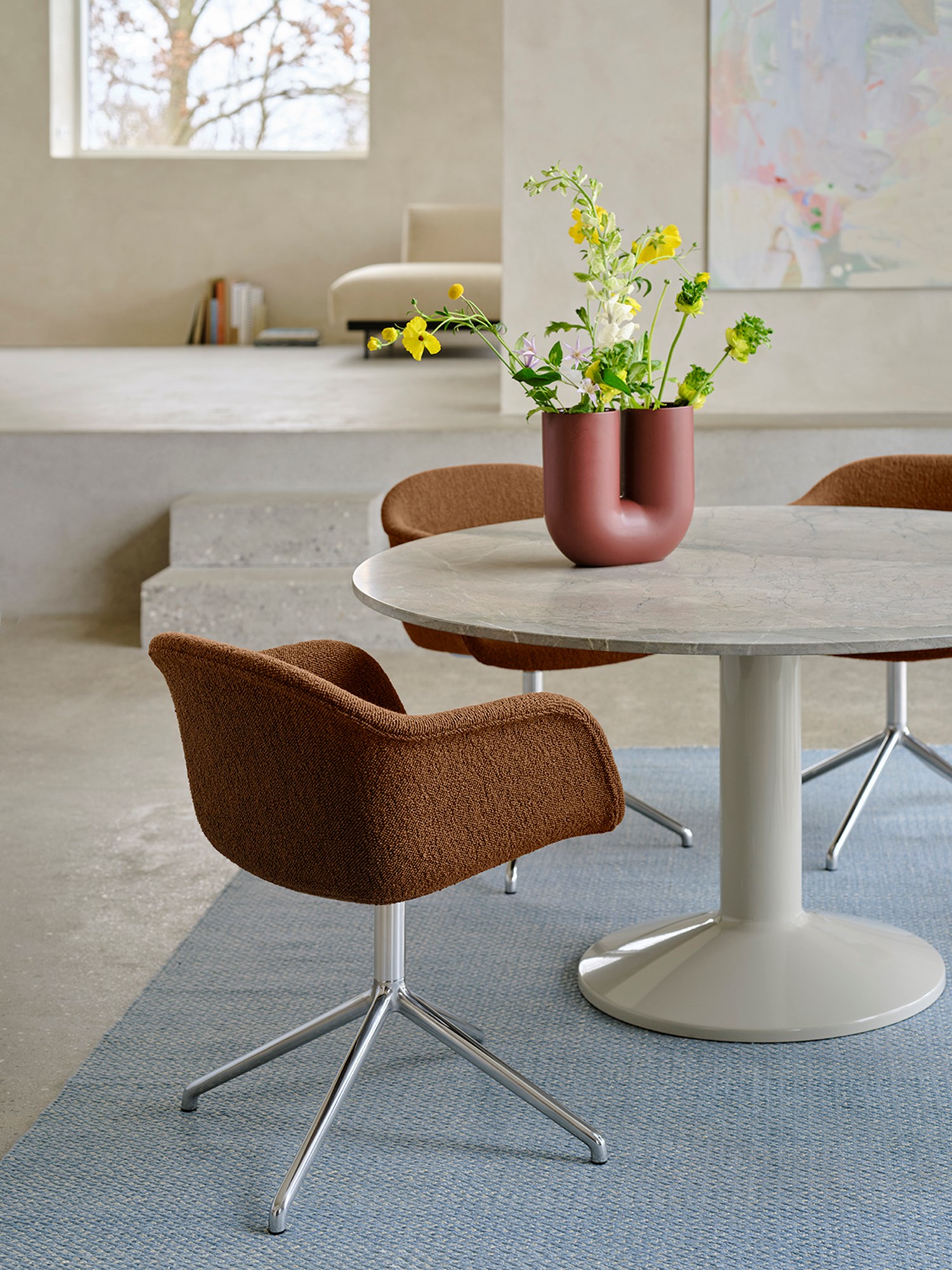 A close-up of a round grey marble dining table in a minimalistic dining room with brushed stone walls and floors. A soft brown dining chair is placed at the table, with a light blue rug underneath adding a pop of color. A sculptural vase with flowers sits on the table, enhancing the serene and modern atmosphere. A close-up of a round grey marble dining table in a minimalistic dining room with brushed stone walls and floors. A soft brown dining chair is placed at the table, with a light blue rug underneath adding a pop of color. A sculptural vase with flowers sits on the table, enhancing the serene and modern atmosphere.