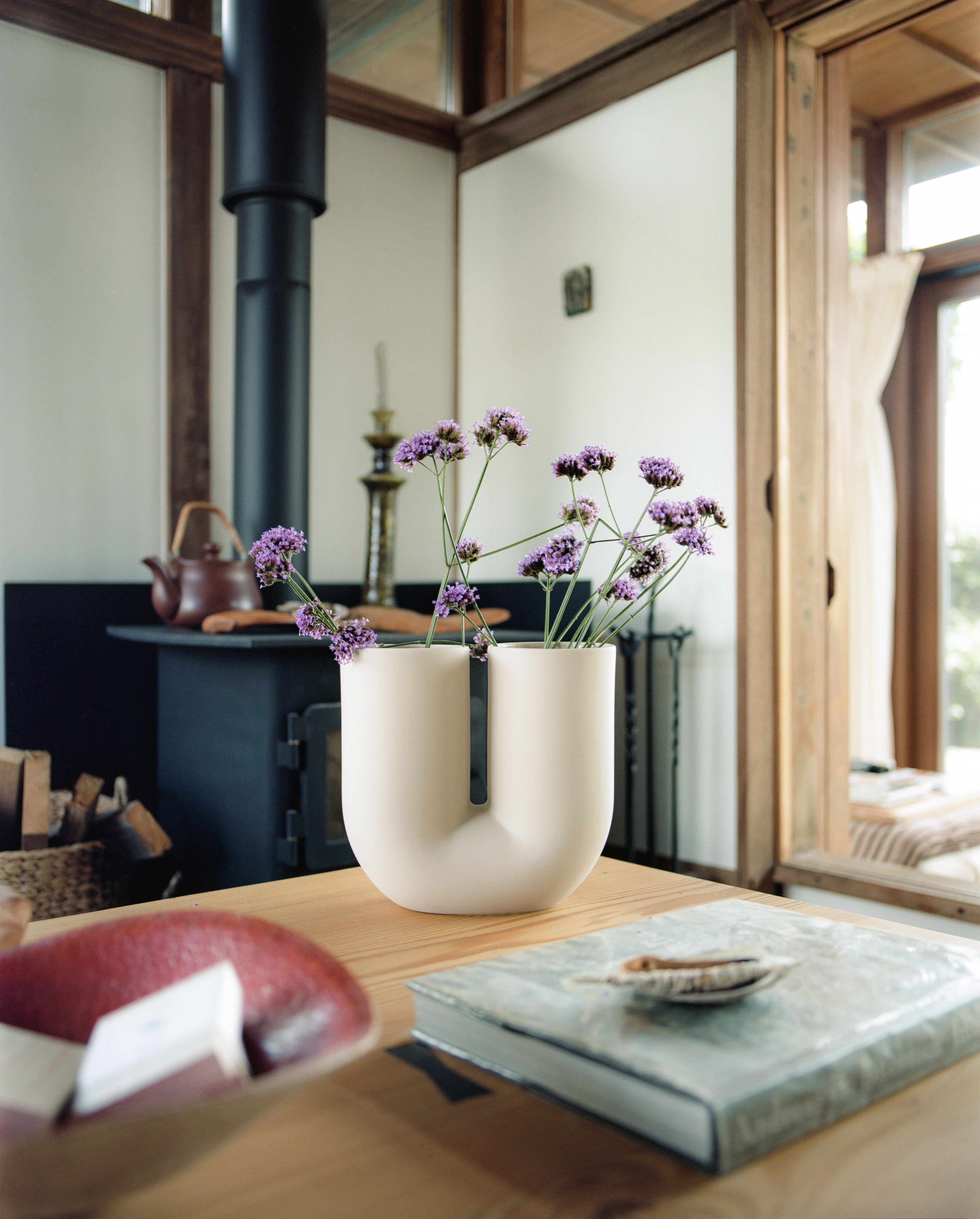 Kink Vase in the color sand with purple flowers on a table in a rustic room with a stove.

