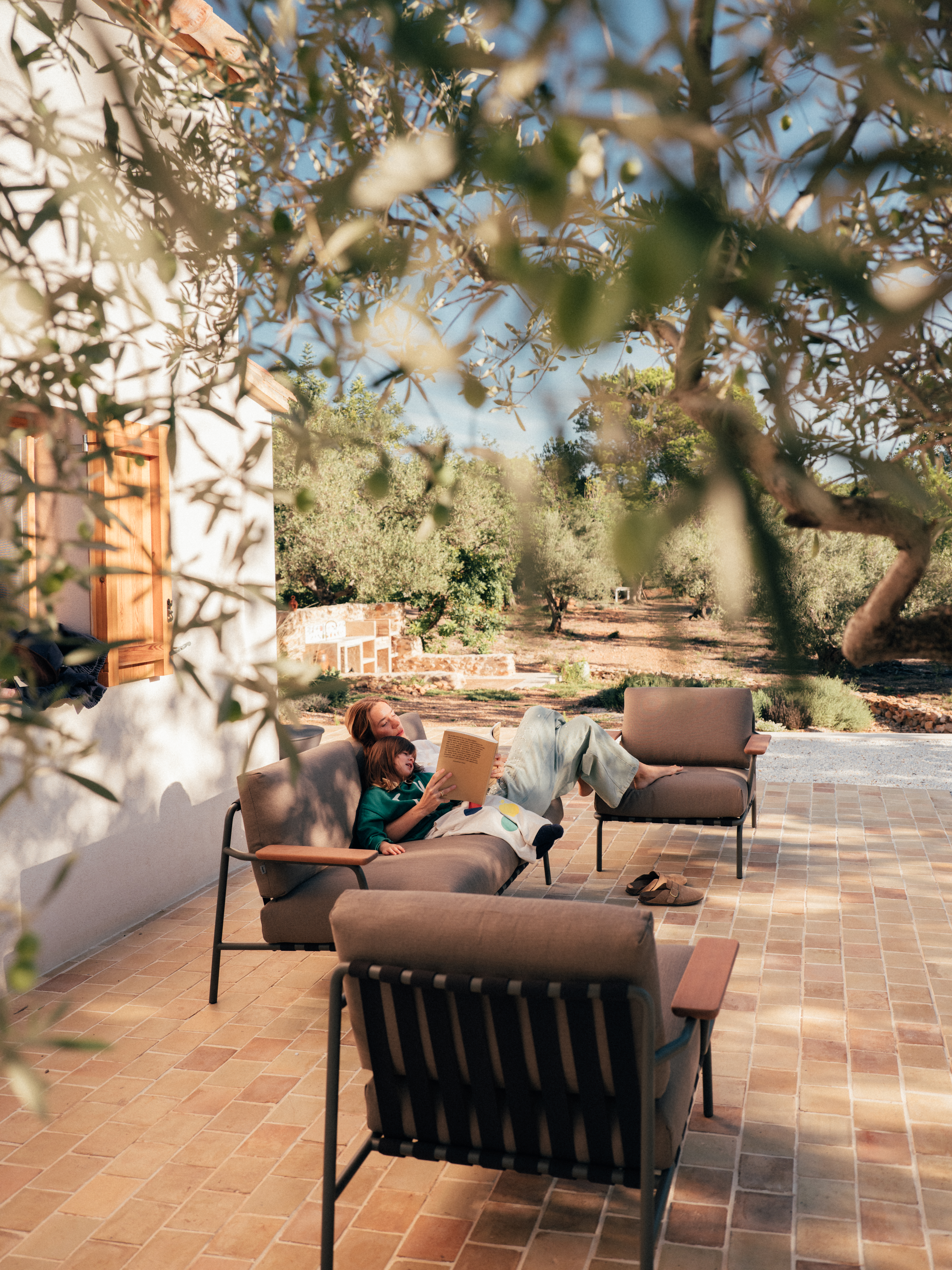 Reading in the afternoon sun – Framed by blurred olive branches, Nuria and her daughter relax on the Settle Sofa on a sunlit patio. She reads aloud from a book, while the child leans against her, nestled in the soft upholstery. The terracotta tiles, warm wooden shutters, and natural surroundings create a tranquil, inviting atmosphere.