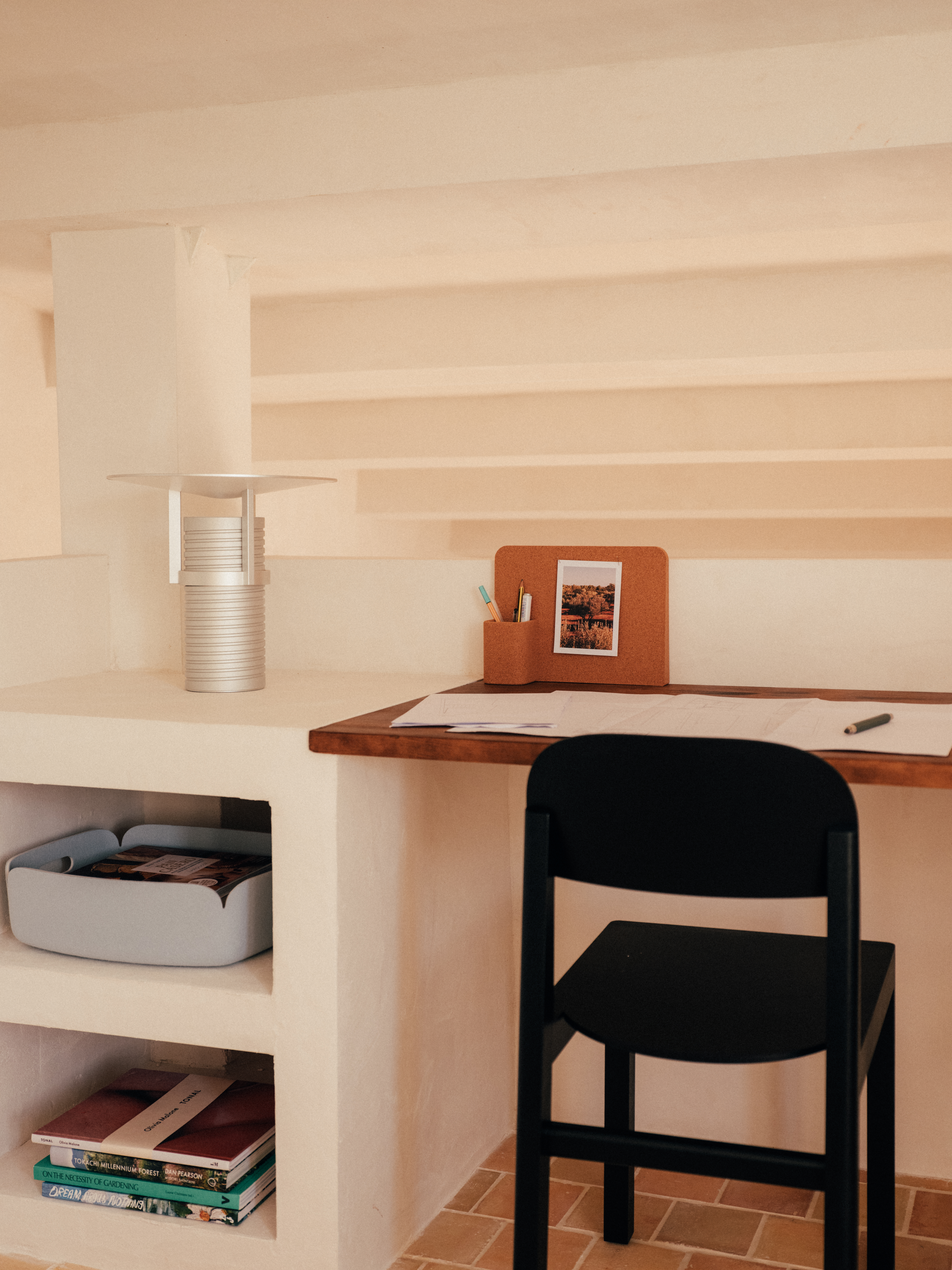 Close-up of a cozy desk setup – A detailed look at a wooden workspace with a sculptural Set Lamp, cork organizer, and a black chair. Below, open built-in shelves store books and a soft gray storage bin, seamlessly blending practicality with the rustic, minimalist decor.