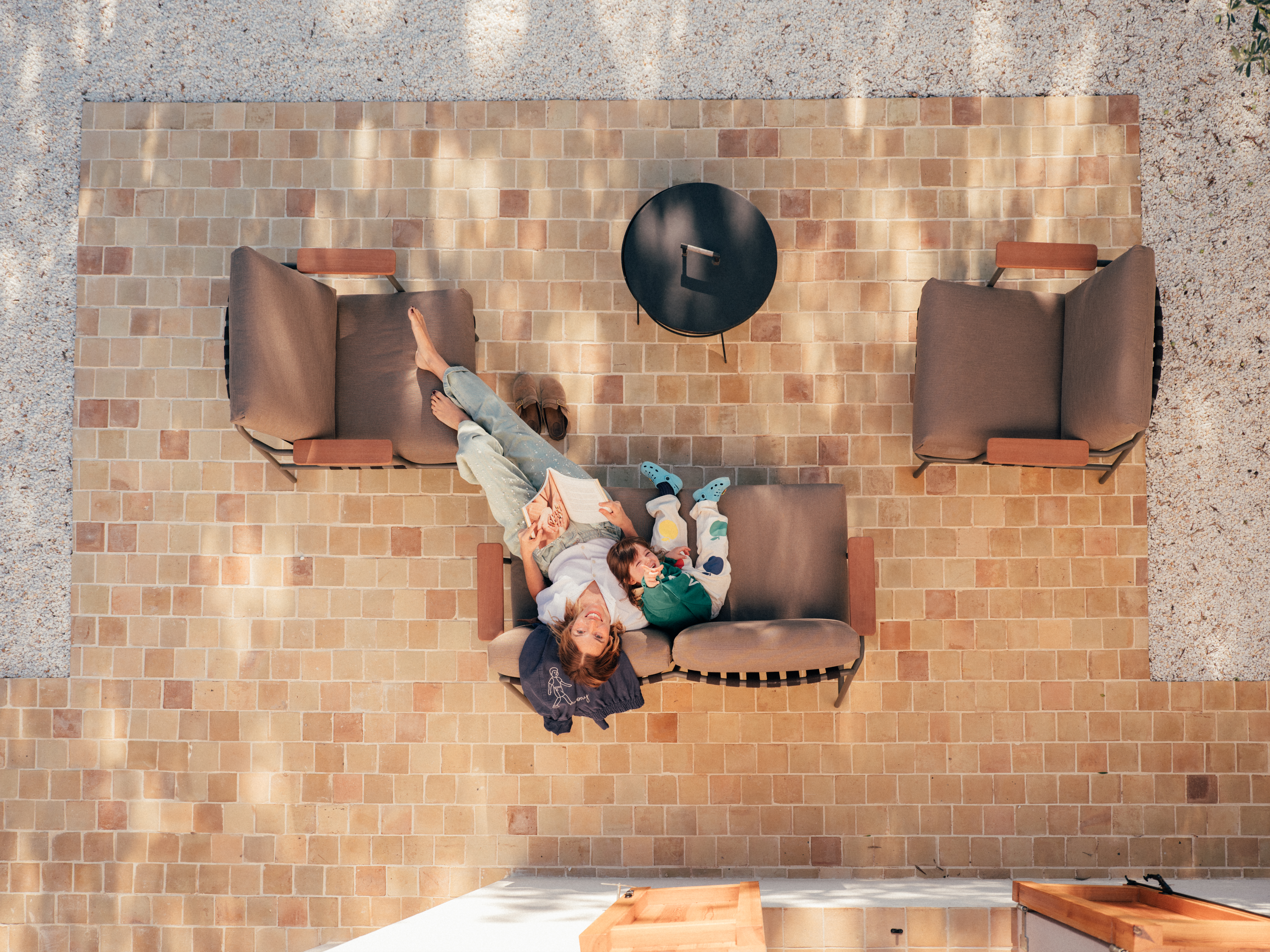 Aerial view of relaxation on a sunlit terrace – From above, Nuria and her daughter lay back on a brown cushioned outdoor sofa. She reads a book while the child leans against her. Two matching armchairs and a fire pit complete the inviting patio, surrounded by soft shadows from the midday sun.