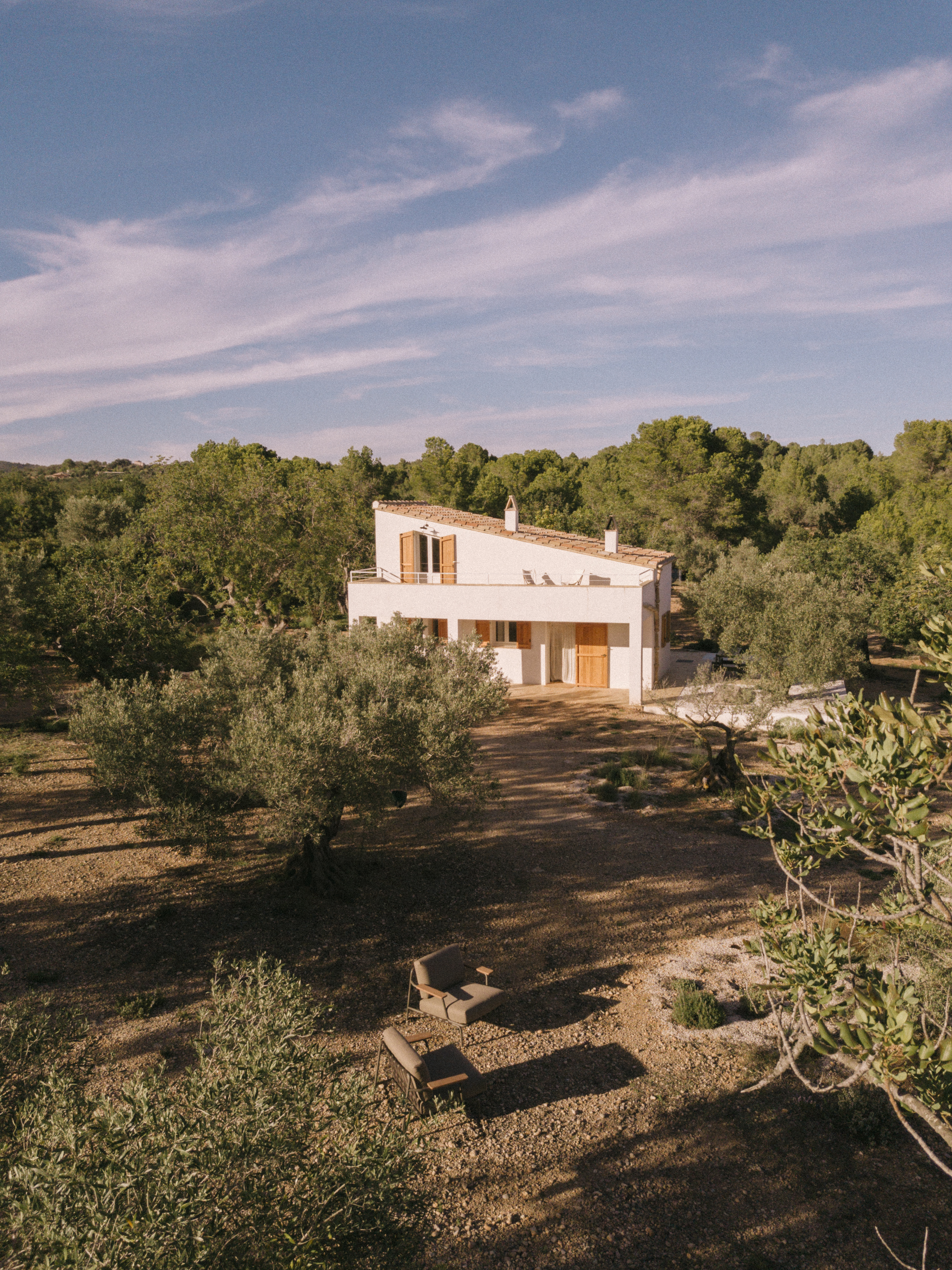 Aerial view of a Mediterranean home – A two-story white stucco house with a terracotta-tiled roof sits among olive trees, bathed in golden light. Wooden shutters and doors accentuate its rustic charm. In the foreground, a cozy outdoor seating area with two brown cushioned chairs and a bench blends seamlessly into the dry, earthy landscape.