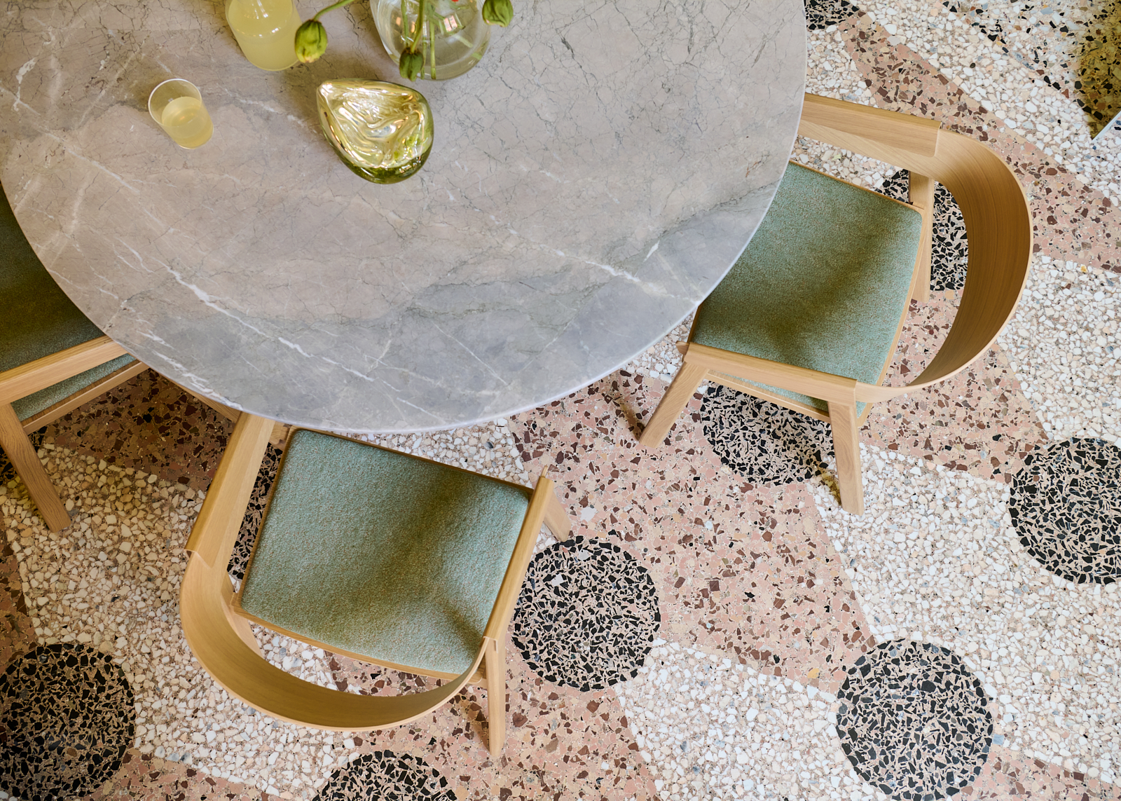 A large marble table seen from above, styled with modern oak dining chairs featuring textile seat pads. The floor has a mosaic pattern that creates a graphic look.