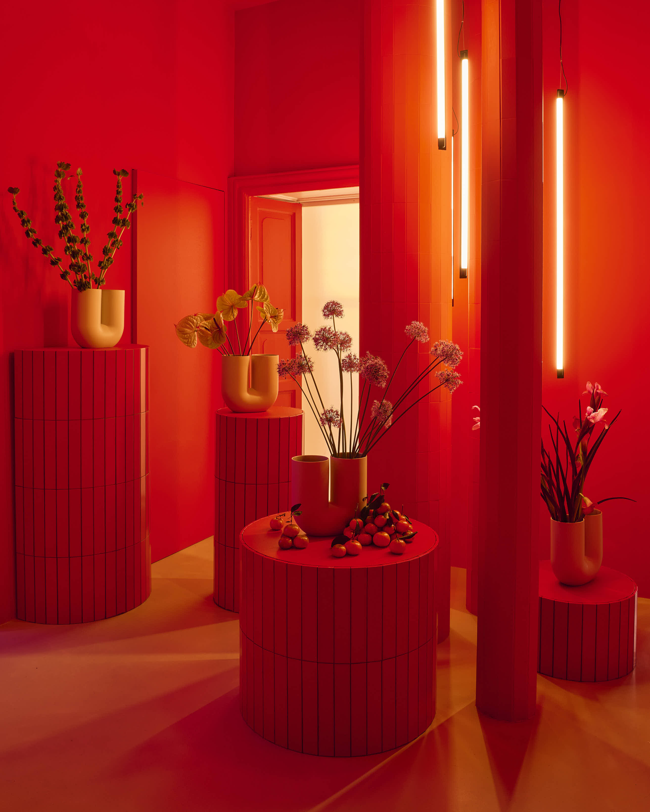 A red room with orange undertones. Pedestals of varying heights are placed around the room, accompanied by sculptural vases with different flowers. 