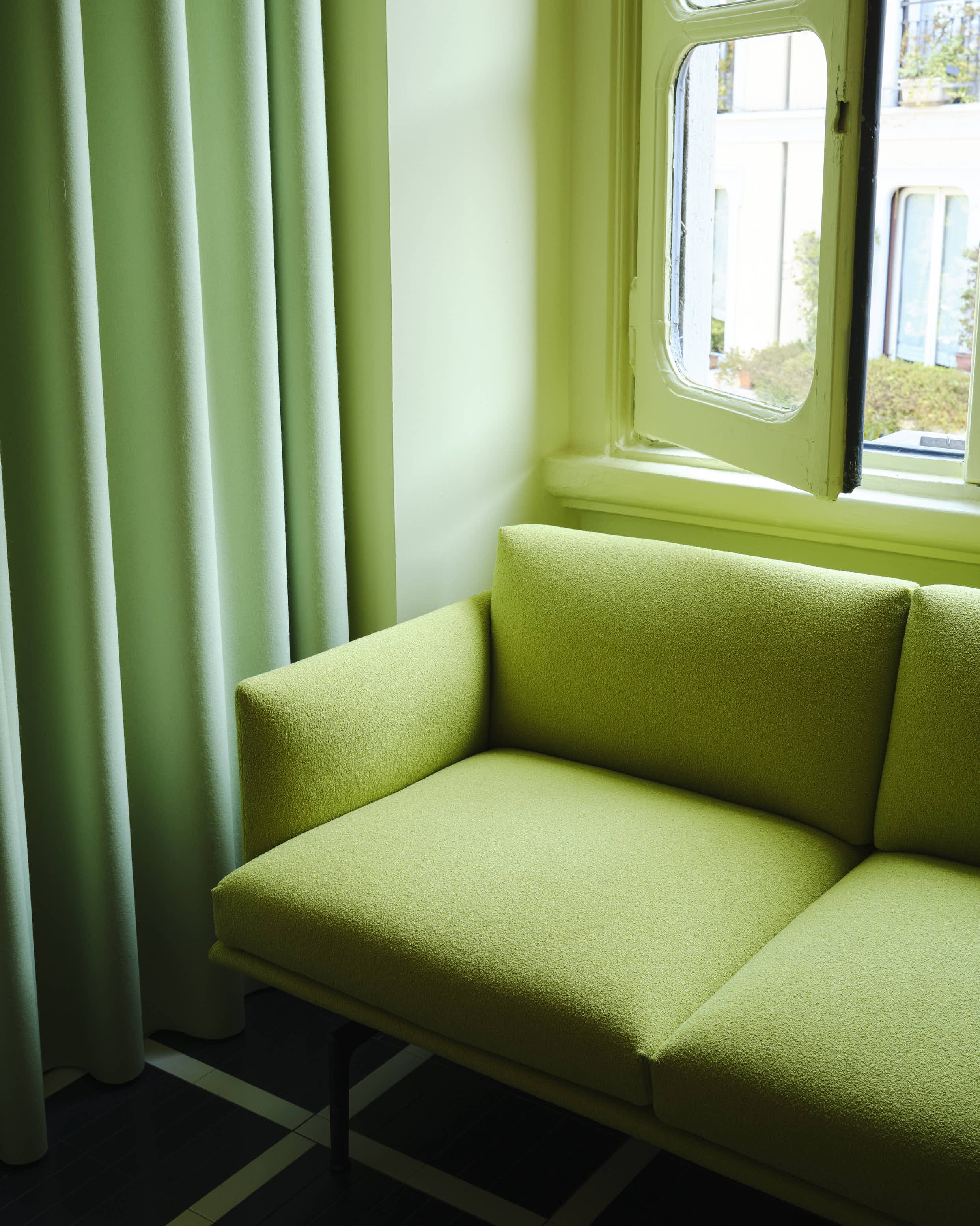 A light green couch is placed in front of an open window. A green curtain hangs beside the couch.