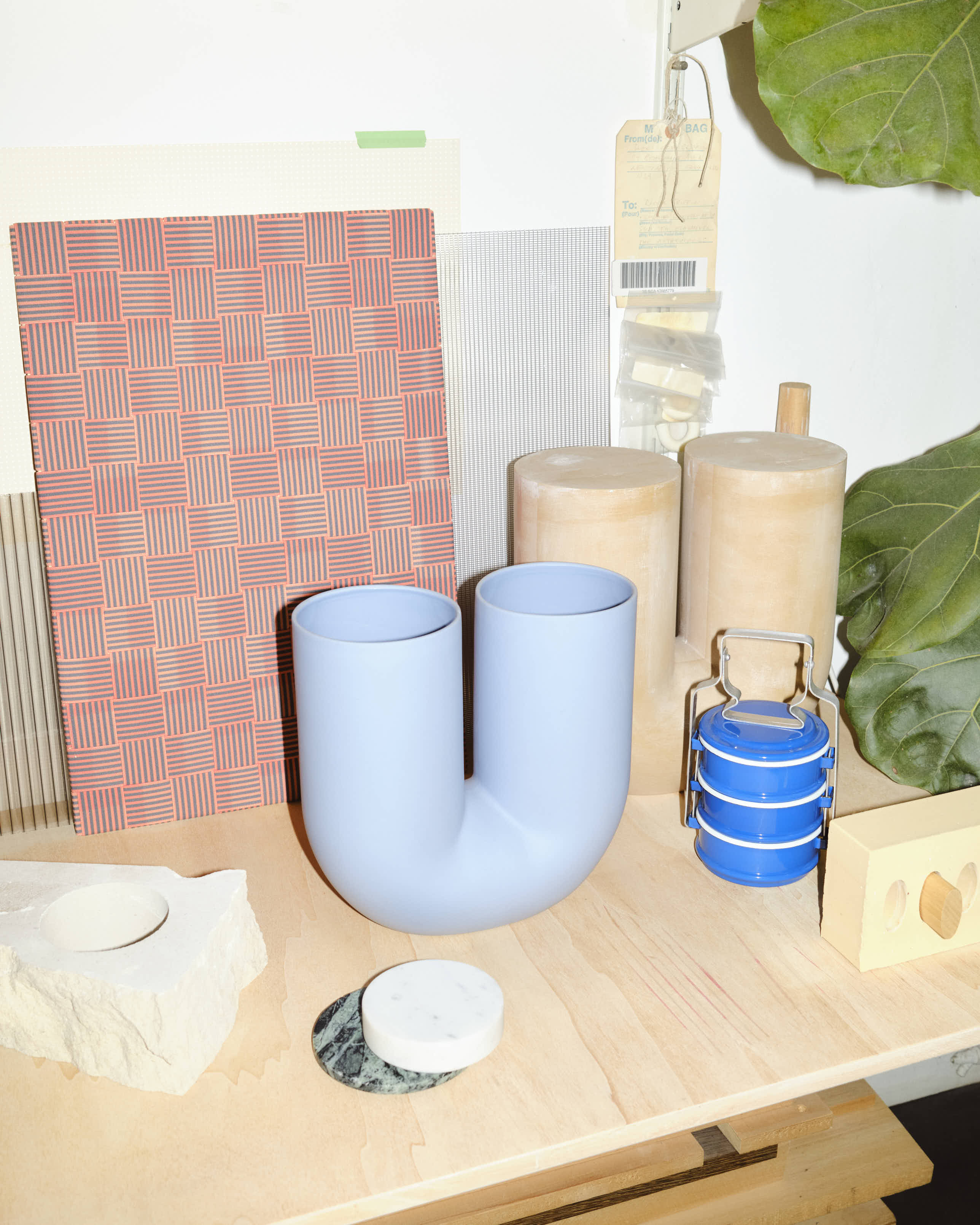 A close-up of the light blue Kink Vase on a wooden surface, surrounded by a textured stone base, circular marble coaster, blue metal tiffin container, and grid-patterned tiles. The vase’s soft matte finish and sculptural form are highlighted by bright lighting.