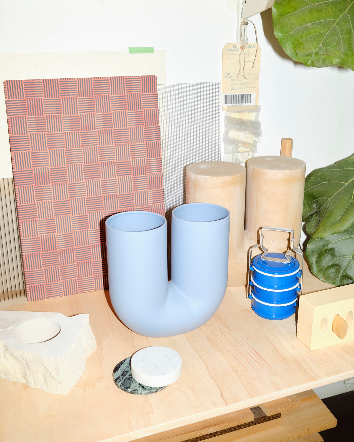 A close-up of the light blue Kink Vase on a wooden surface, surrounded by a textured stone base, circular marble coaster, blue metal tiffin container, and grid-patterned tiles. The vase’s soft matte finish and sculptural form are highlighted by bright lighting. A close-up of the light blue Kink Vase on a wooden surface, surrounded by a textured stone base, circular marble coaster, blue metal tiffin container, and grid-patterned tiles. The vase’s soft matte finish and sculptural form are highlighted by bright lighting.