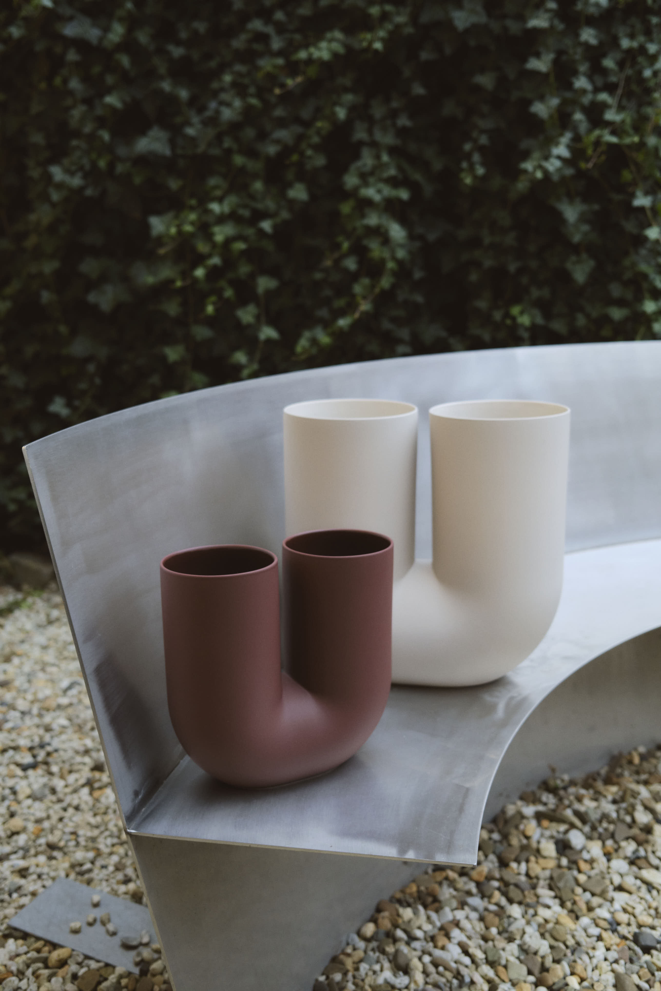 Two Kink Vases—one in deeo red, the other in creamy white—sit side-by-side on a curved metal bench in an outdoor garden setting. The gravel ground and leafy green backdrop contrast with the vases’ smooth, modern ceramic forms.