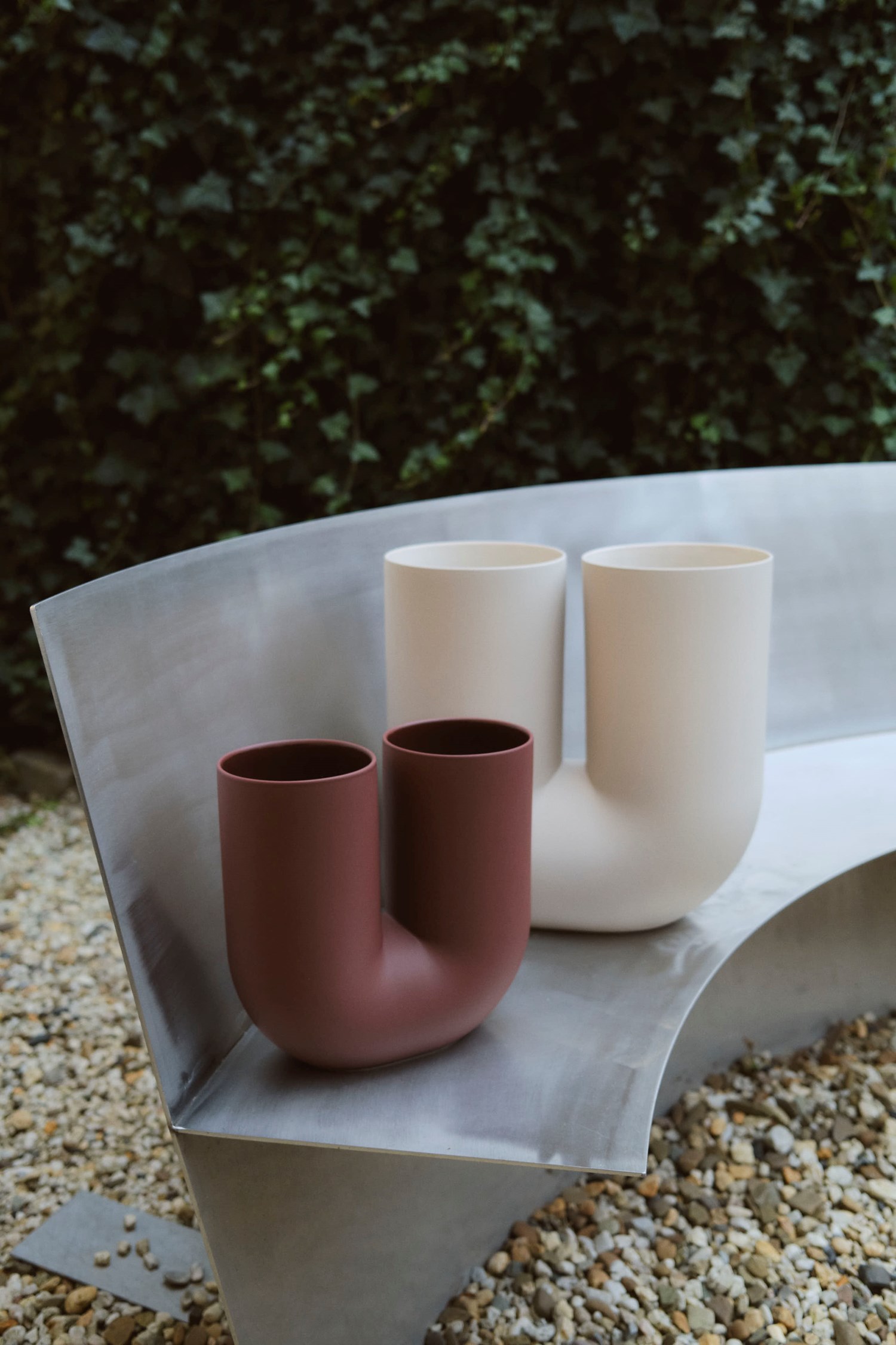 Two Kink Vases—one in deeo red, the other in creamy white—sit side-by-side on a curved metal bench in an outdoor garden setting. The gravel ground and leafy green backdrop contrast with the vases’ smooth, modern ceramic forms. Two Kink Vases—one in deeo red, the other in creamy white—sit side-by-side on a curved metal bench in an outdoor garden setting. The gravel ground and leafy green backdrop contrast with the vases’ smooth, modern ceramic forms.