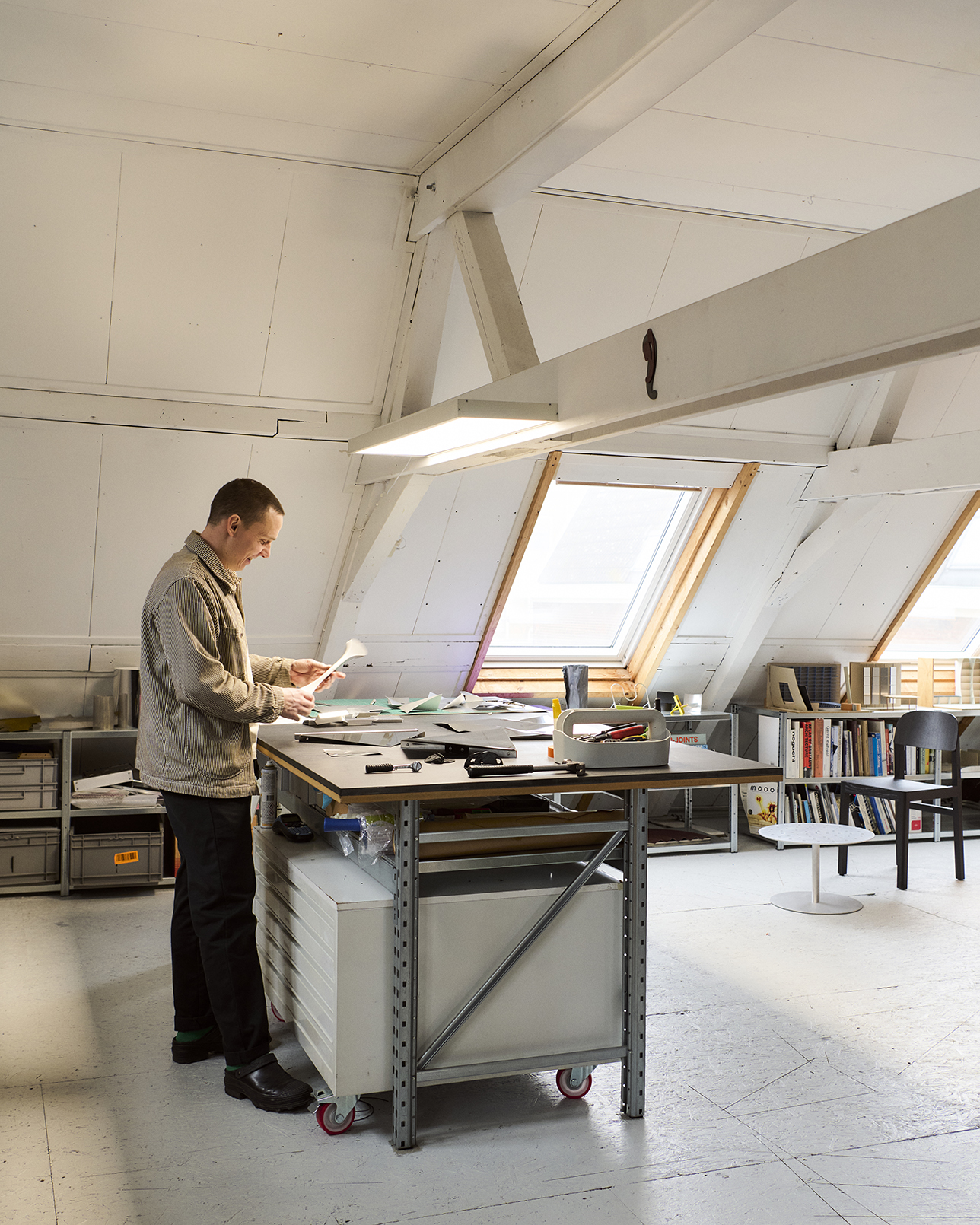 In a bright attic studio with slanted skylights and exposed beams, Phil stands at a central metal-framed workbench examining paper or fabric samples.


