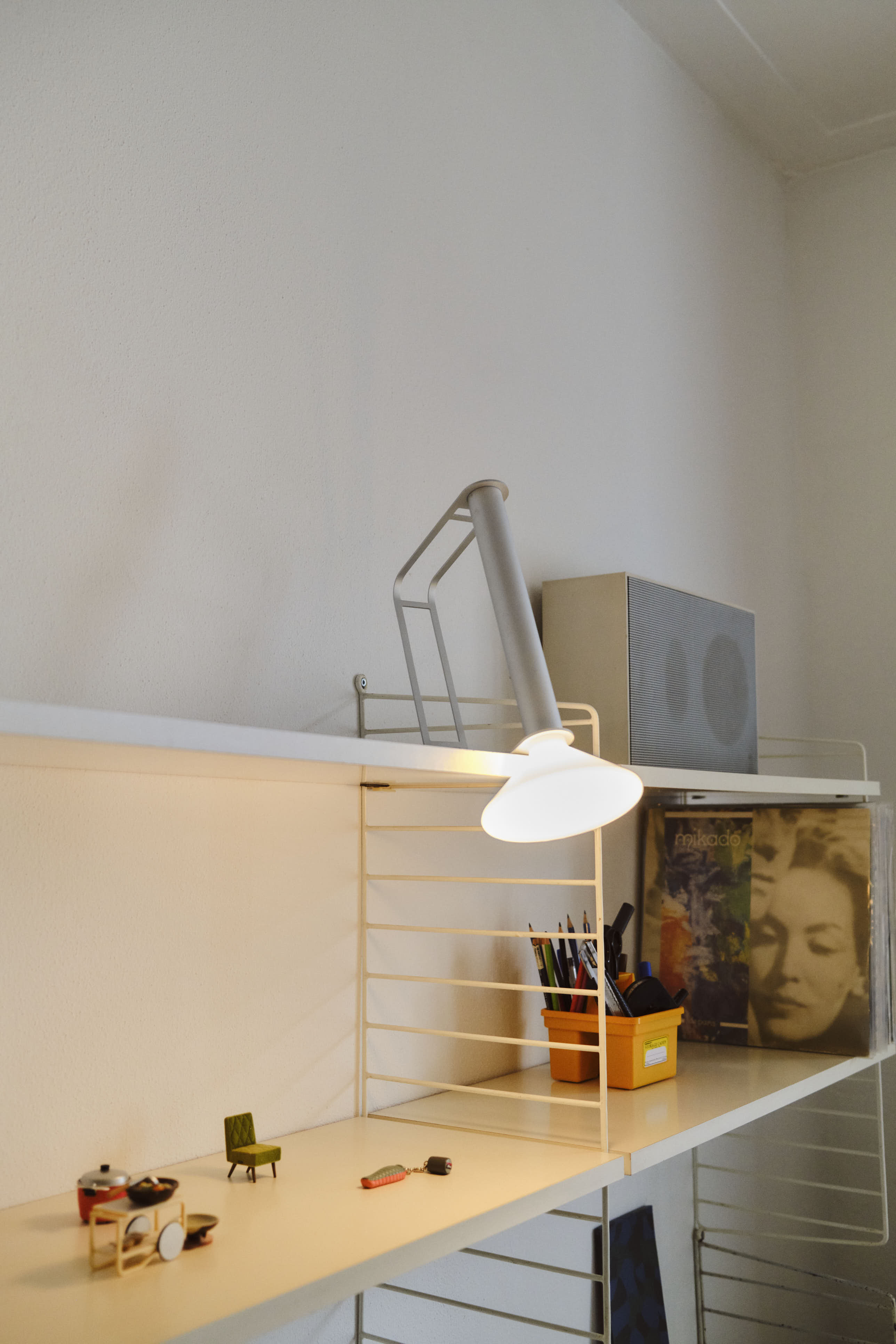 A modern white metal shelf featuring a silver Piton Portable Lamp casting a warm glow. The shelf holds small decorative items, a container of pens, and a collection of vinyl records, creating a stylish and functional workspace.