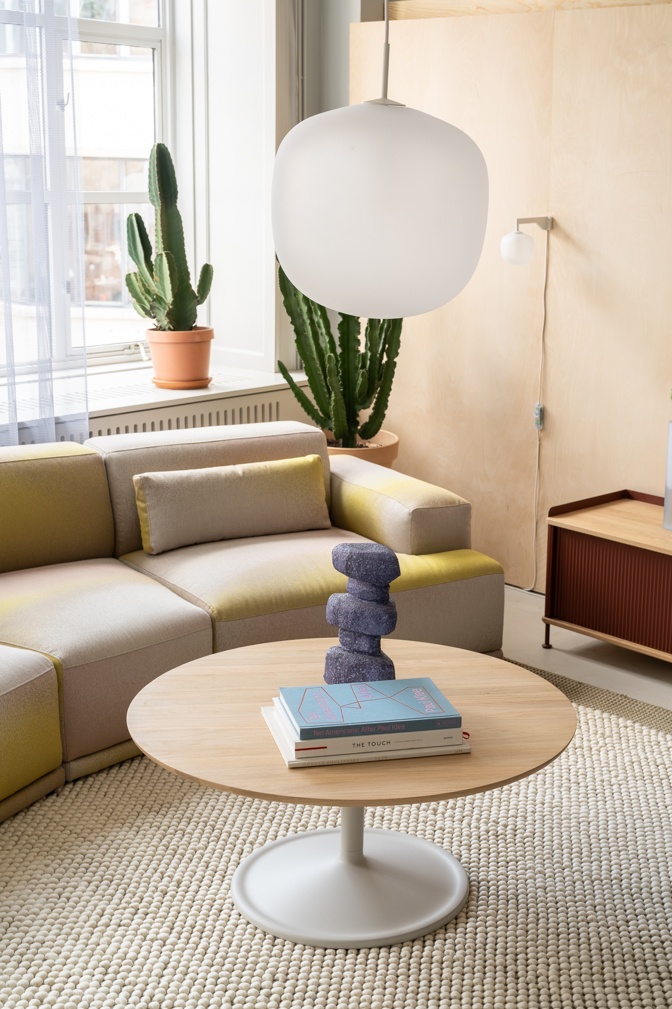 A round Soft Coffee Table is placed on a beige rug in a simple - yet modern - lounge area. A pendant lamp is hanging from the ceiling and a colored sofa is placed in the room creating a comfy feeling. 