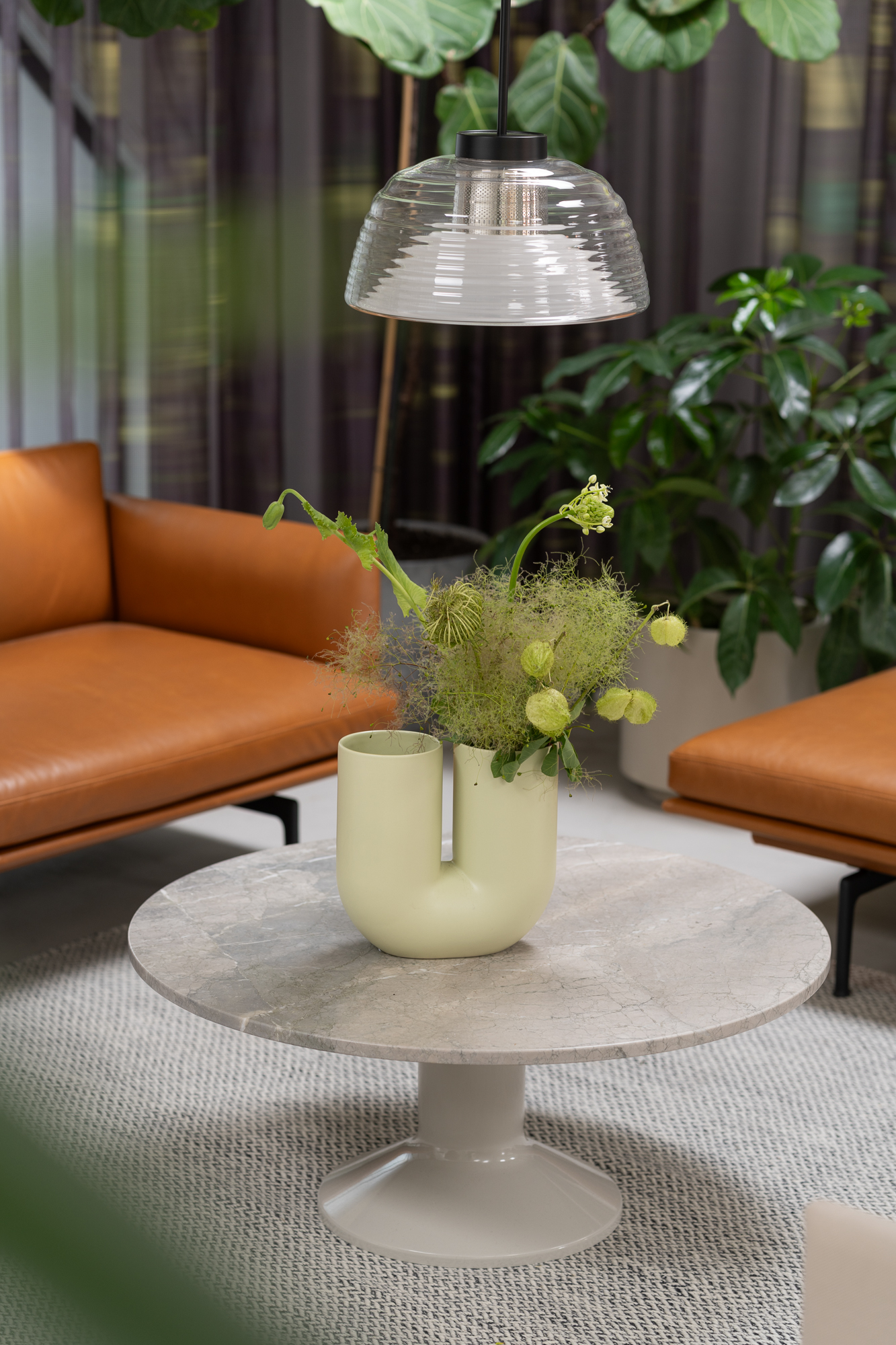 A Midst Coffee Table is placed on a grey rug surrounded by brown leather sofas. A sculptural vase with green plants and flowers is placed on the table. 