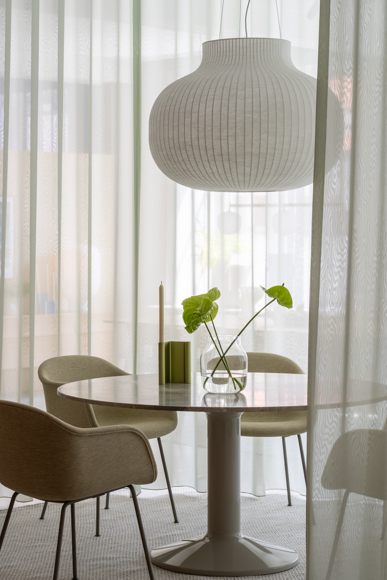 The Midst table in marble is placed in a white room with modern curtains. Soft green chairs surround the table, while a pendant lamp hangs above, adding a cozy touch to the space.