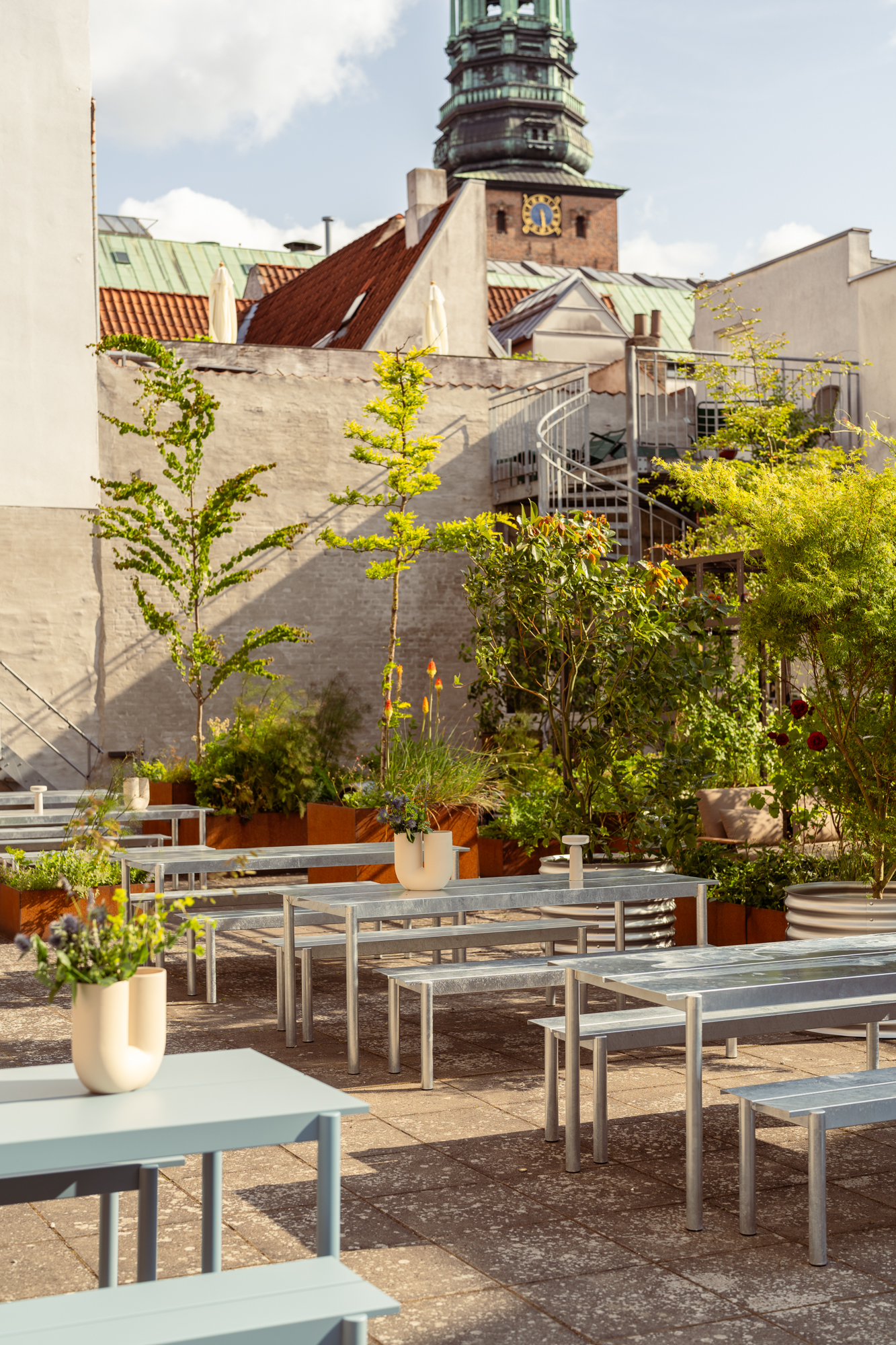 Linear Steel tables and benches in light blue are arranged in two rows in a cozy backyard surrounded by green trees and lush plants. The artistic Kink Vase sits atop the tables, styled with a creative floral arrangement that adds a sculptural touch.