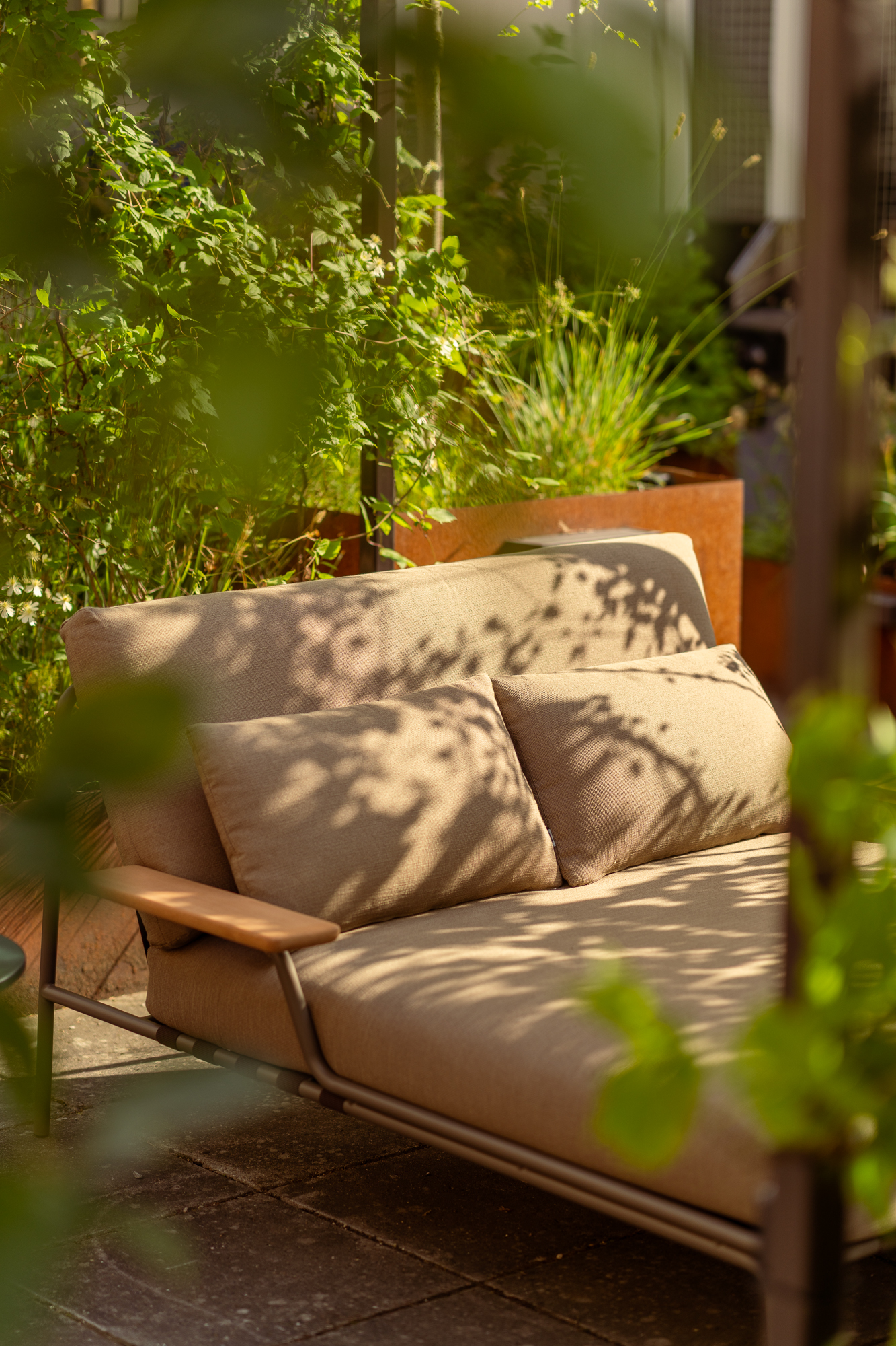 A Settle lounge bed with armrests is placed in a sunny courtyard, surrounded by lush green plants. 