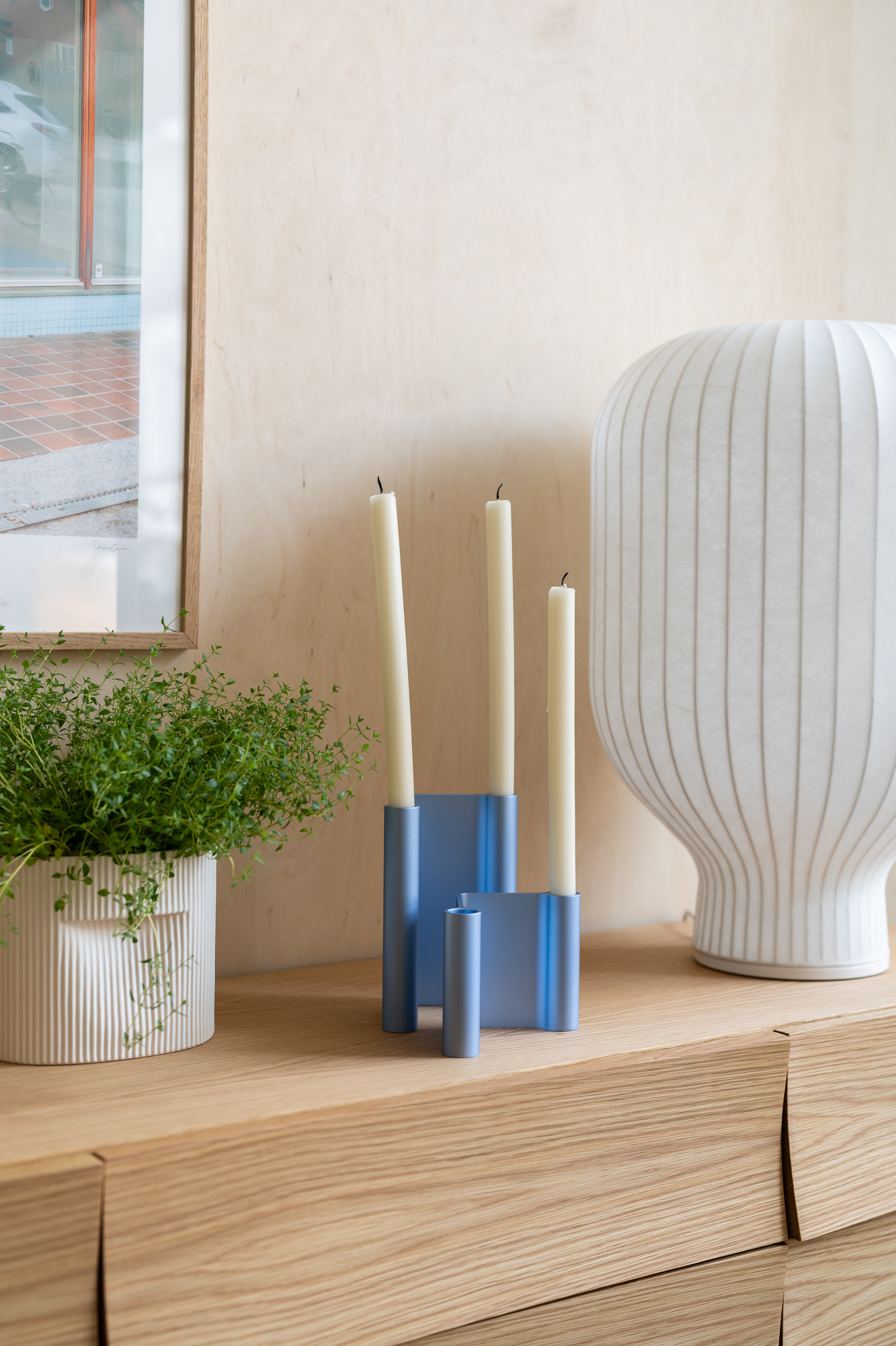 The Strand Lamp sits elegantly on a wooden sideboard, accompanied by a blue candle holder and a white vase containing a green plant. The arrangement creates a simple yet stylish vignette with a natural touch.