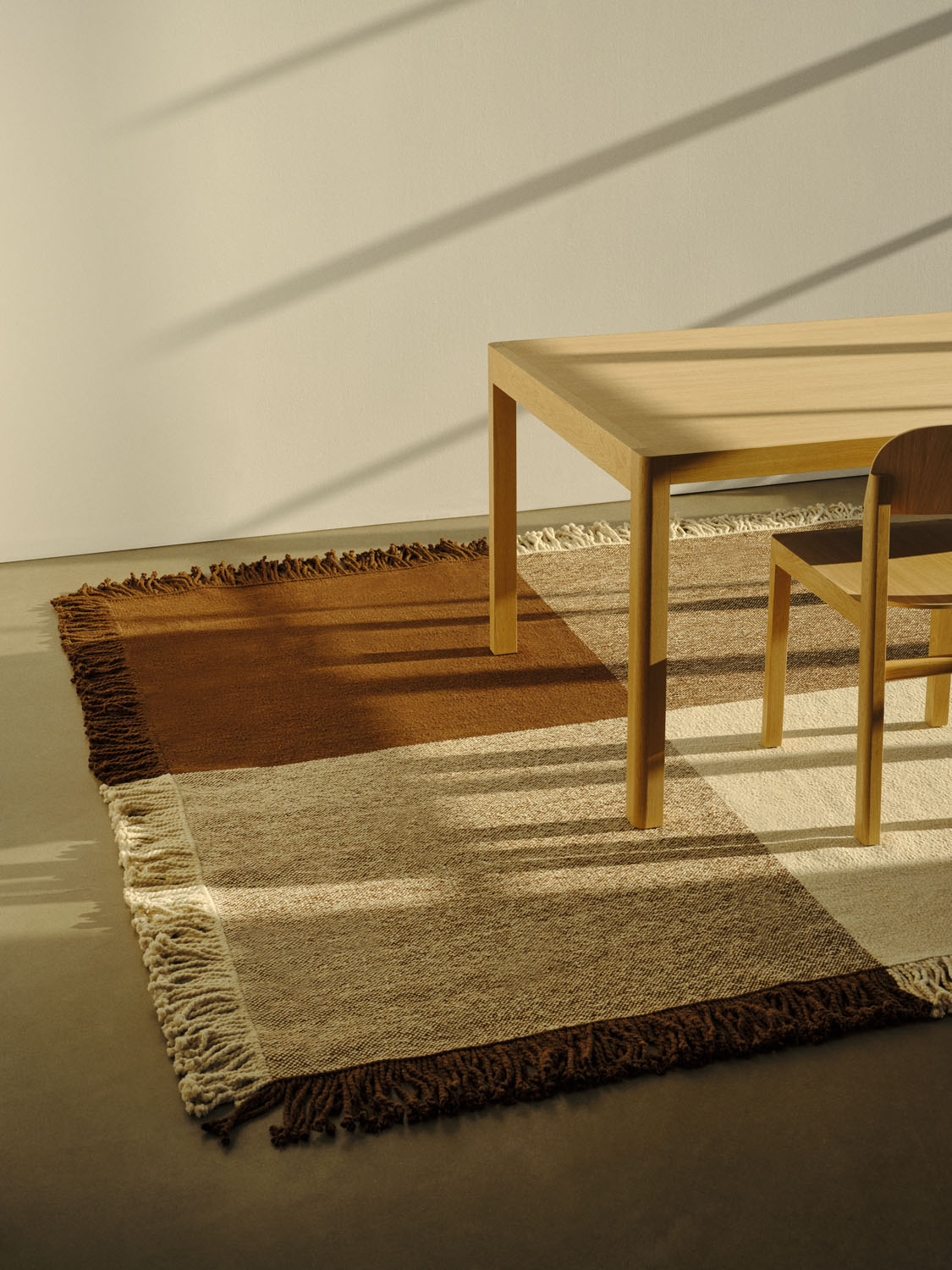 A clean, modern interior featuring the Omni rug beneath a wooden table and chair. The rug’s color-blocked pattern in rust brown, speckled tan, and light beige adds warmth to the neutral space. Afternoon light casts long shadows across the minimalist setting, highlighting the handwoven detail.