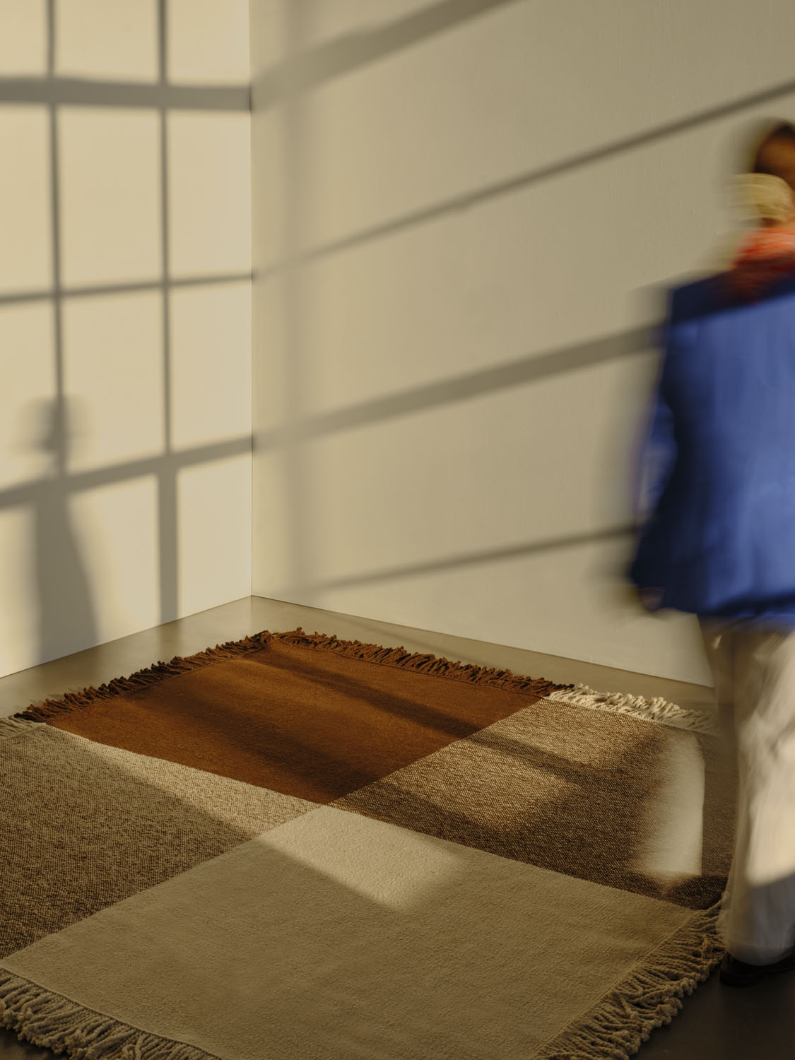 A full view of Sia’s Omni rug laid out on a minimalist floor, with a person walking past in motion blur. The rug showcases a geometric patchwork of brown, beige, and cream squares, bordered by dark and light fringe. The soft, angled sunlight creates dramatic shadows across the walls and rug, emphasizing its rich texture.