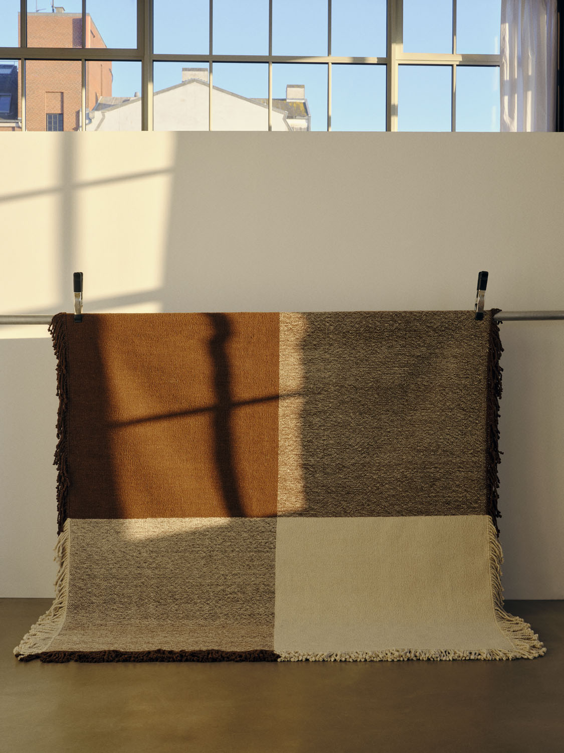 The Omni rug is clipped to a rail in a bright, loft-like space with large windows. The rug’s rich brown and tan blocks and dense fringe are illuminated by natural light, casting windowpane shadows across its textured surface.
