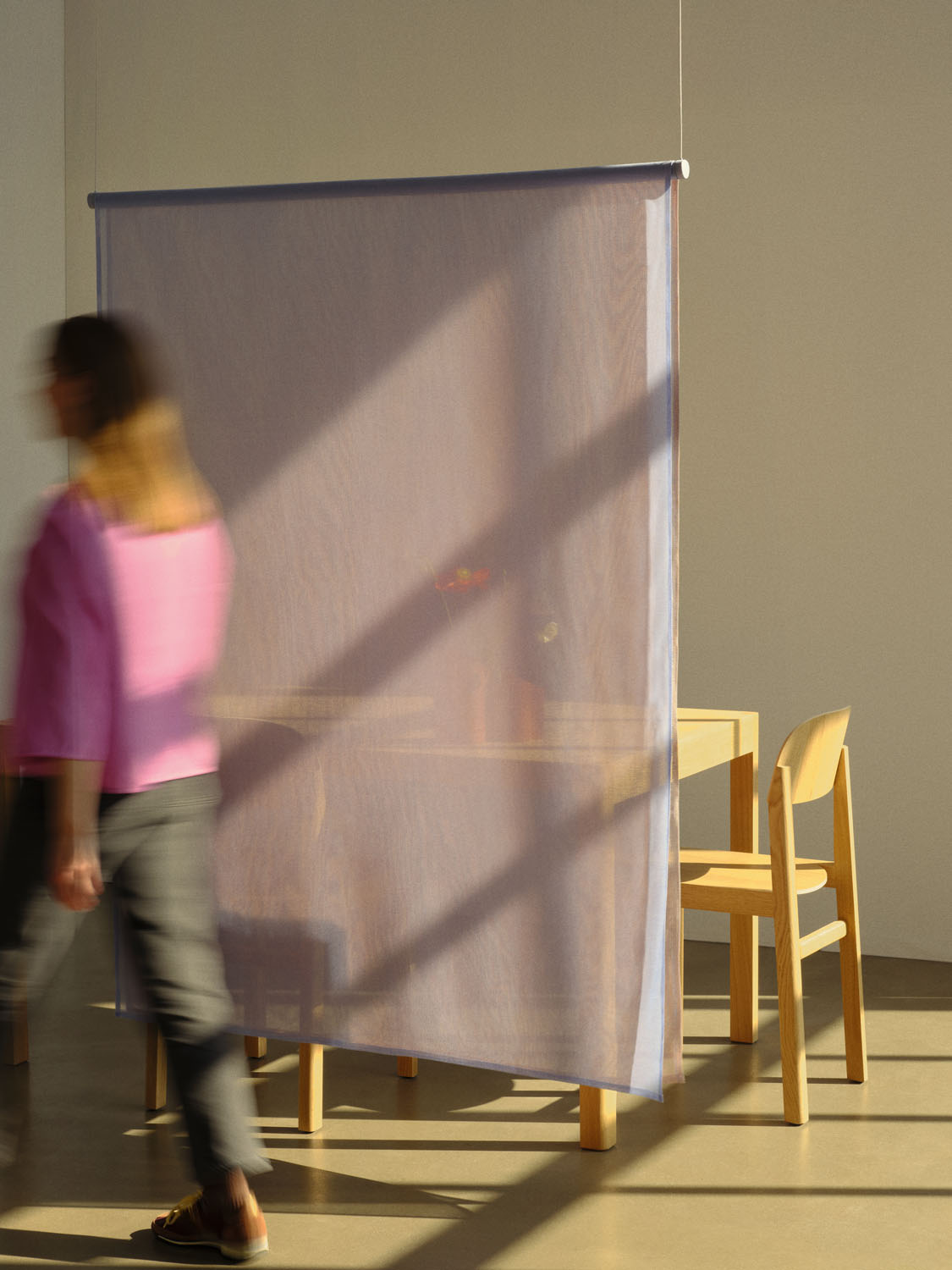 A translucent lavender mesh panel from the “Veil” collection hangs in a softly lit space, partially concealing a dining table and wooden chairs behind it. A person in motion, dressed in a pink top and gray pants, walks by, creating a blurred silhouette and evoking a sense of privacy in motion.

