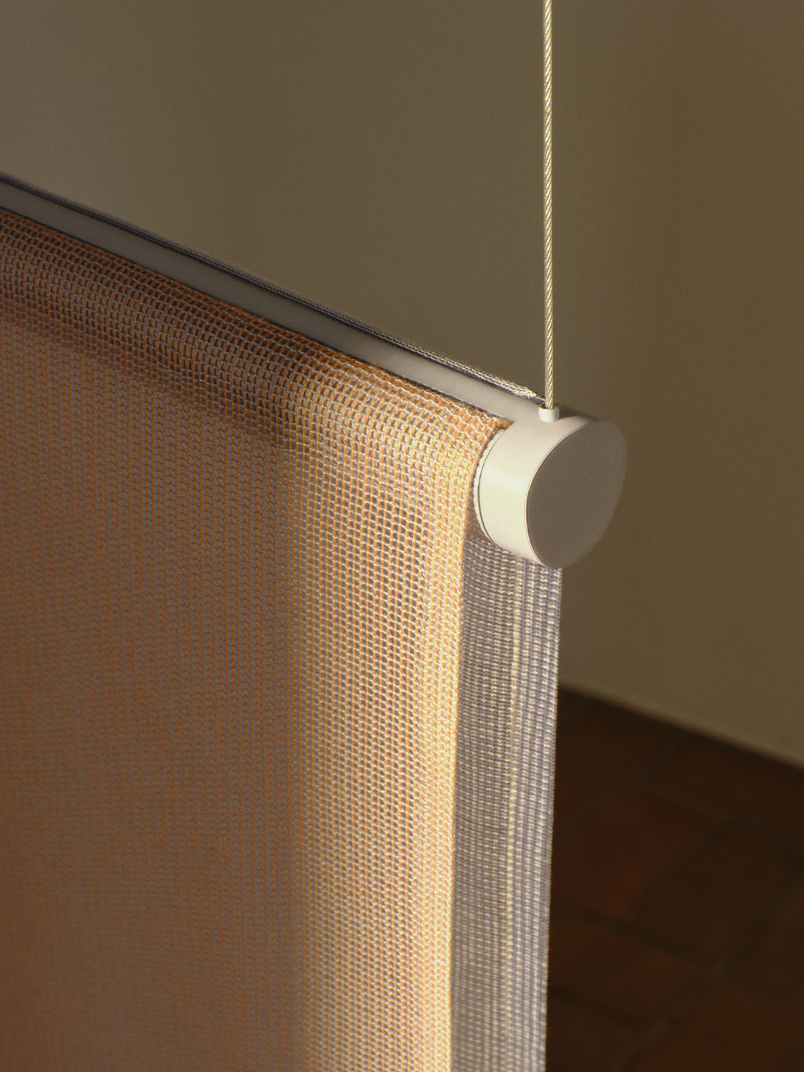 Detail of the top corner of a suspended “Veil” fabric panel in soft orange mesh. The panel is attached to a cylindrical aluminum track, suspended by a thin steel wire. 