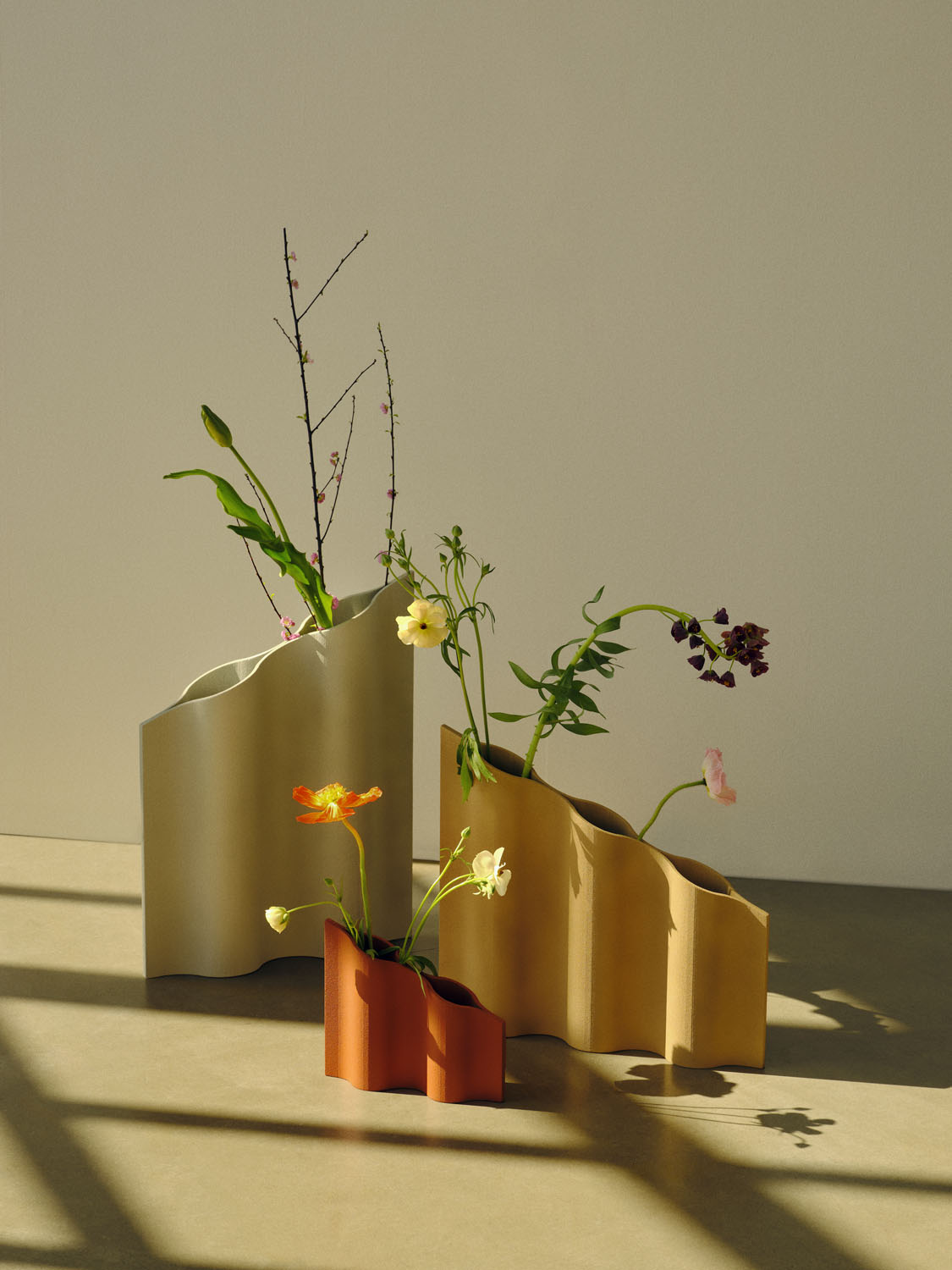 Three sculptural “Waves” prototype vases by Francesc Gasch, with undulating, rhythmic silhouettes. Each vase has a stepped wave-like profile and is displayed on a concrete floor in natural sunlight. The tallest vase is a pale cream, the medium one is ochre, and the smallest is terracotta red. Each holds a minimal floral arrangement.

