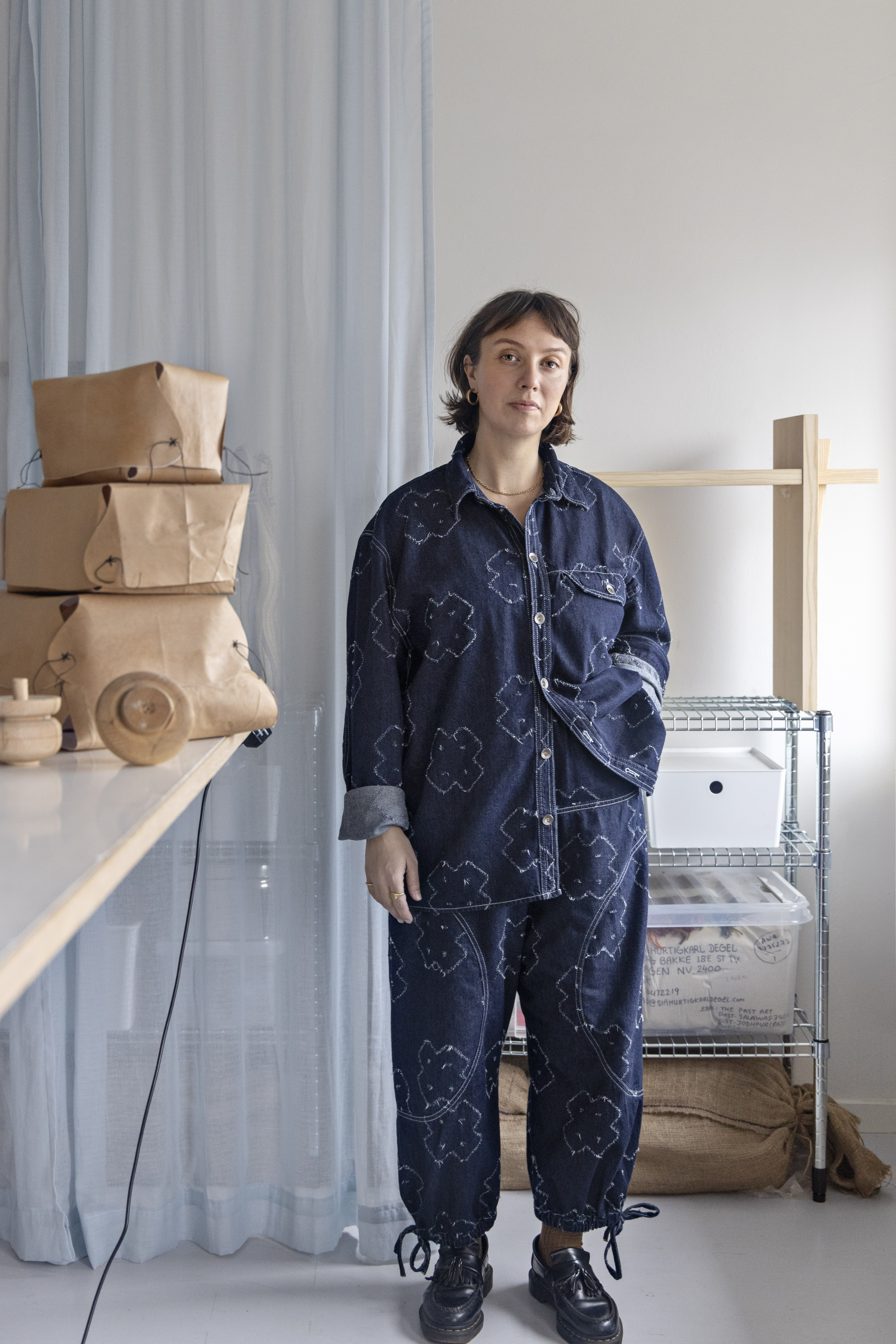 Portrait of Danish designer Sia standing in her studio beside shelves and paper prototypes. She wears a dark blue matching set with stitched motifs, embodying a functional and creative style. The space around her features soft lighting, stacked kraft paper models.