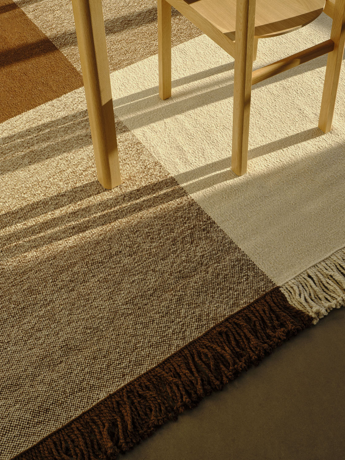 Close-up of the Omni rug beneath a wooden chair leg. The intricate weave in cream, warm brown, and tan tones, along with dark brown fringe, is captured in golden sunlight. The rug’s soft texture and bold grid pattern add natural elegance to the space.
