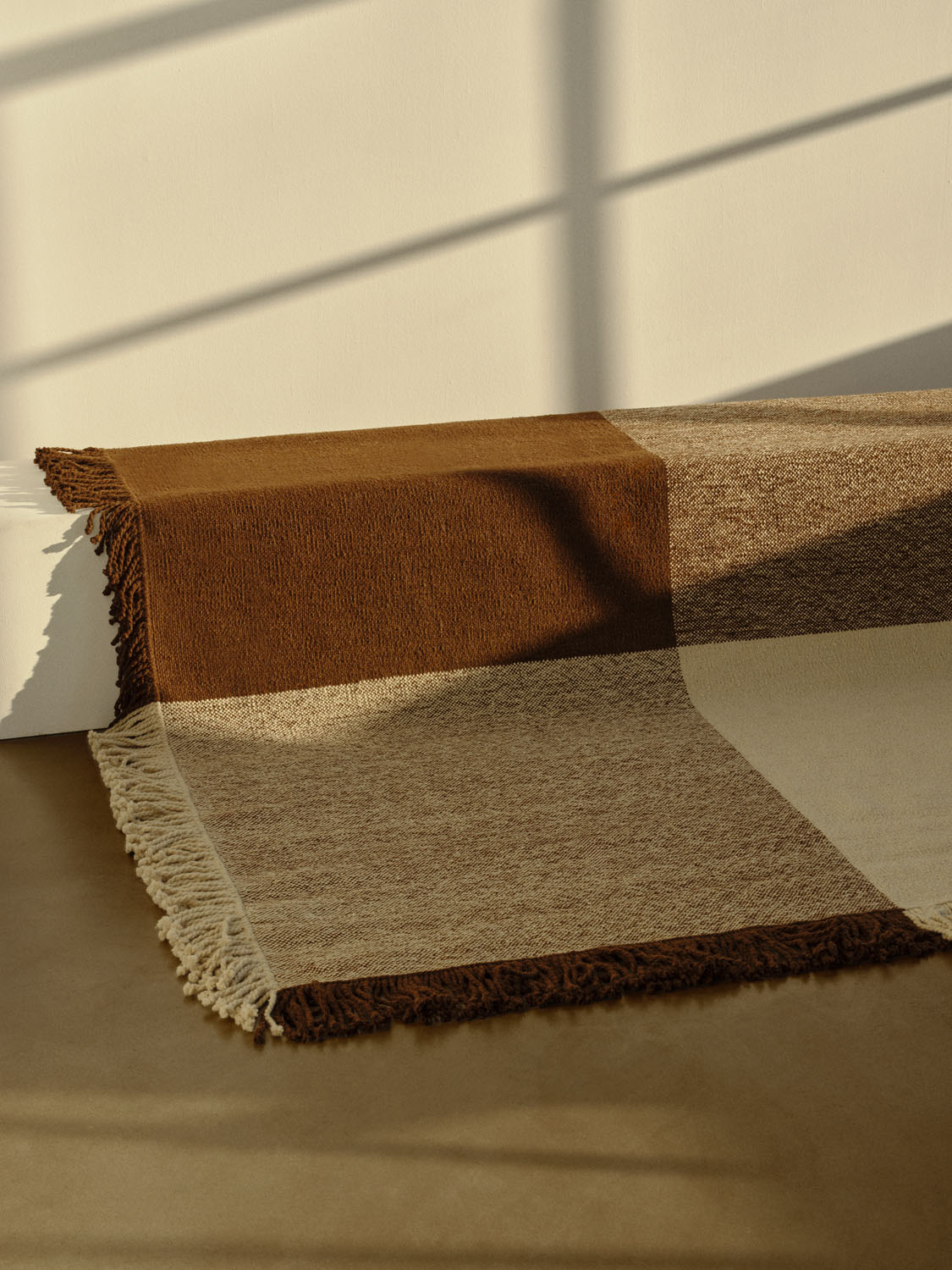The Omni rug by Sia draped over a low ledge in a sunlit room. The design features a large-scale block pattern in warm browns, tan, and cream, woven with visible texture. Thick fringes border the edges, and sunlight casts soft shadows across the neutral-toned floor and wall.


