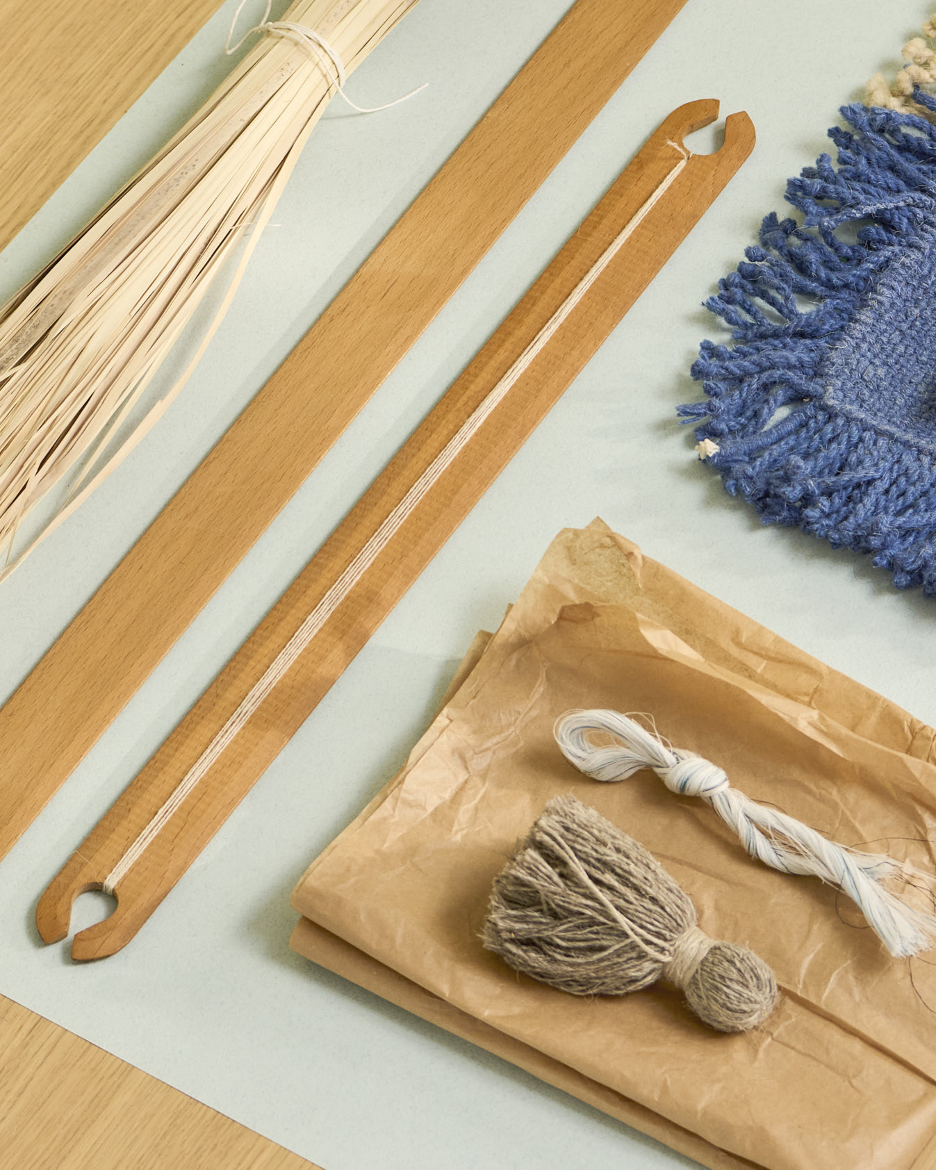 A flat lay of weaving tools and natural materials used in the making of the Omni rug. The composition includes two slender wooden weaving shuttles, a wrapped bundle of straw-like plant fibers, skeins of rough and smooth yarn in beige and white, and a vibrant blue woven textile sample with thick fringe. The items rest on soft paper and a pale background, evoking a calm, tactile workspace rooted in craft and natural fibers.