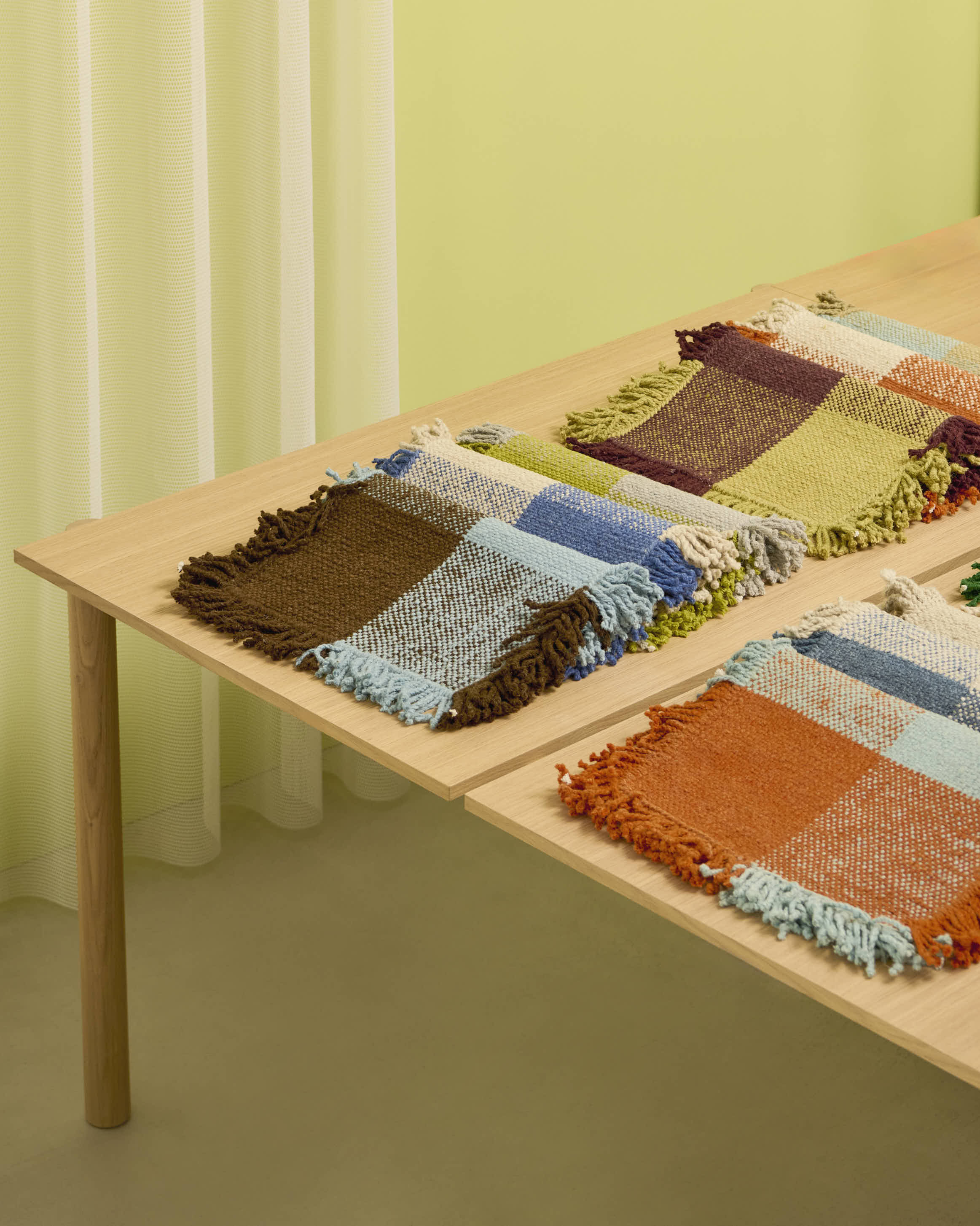 Close-up of Omni rug samples displayed on a pale wood surface. The foreground features a swatch in rich brown, sky blue, and cream tones with textured fringes, while additional swatches in olive green, navy, and mustard hues are visible. The backdrop is a pale green wall and sheer white curtain, creating a warm, modern showroom setting.