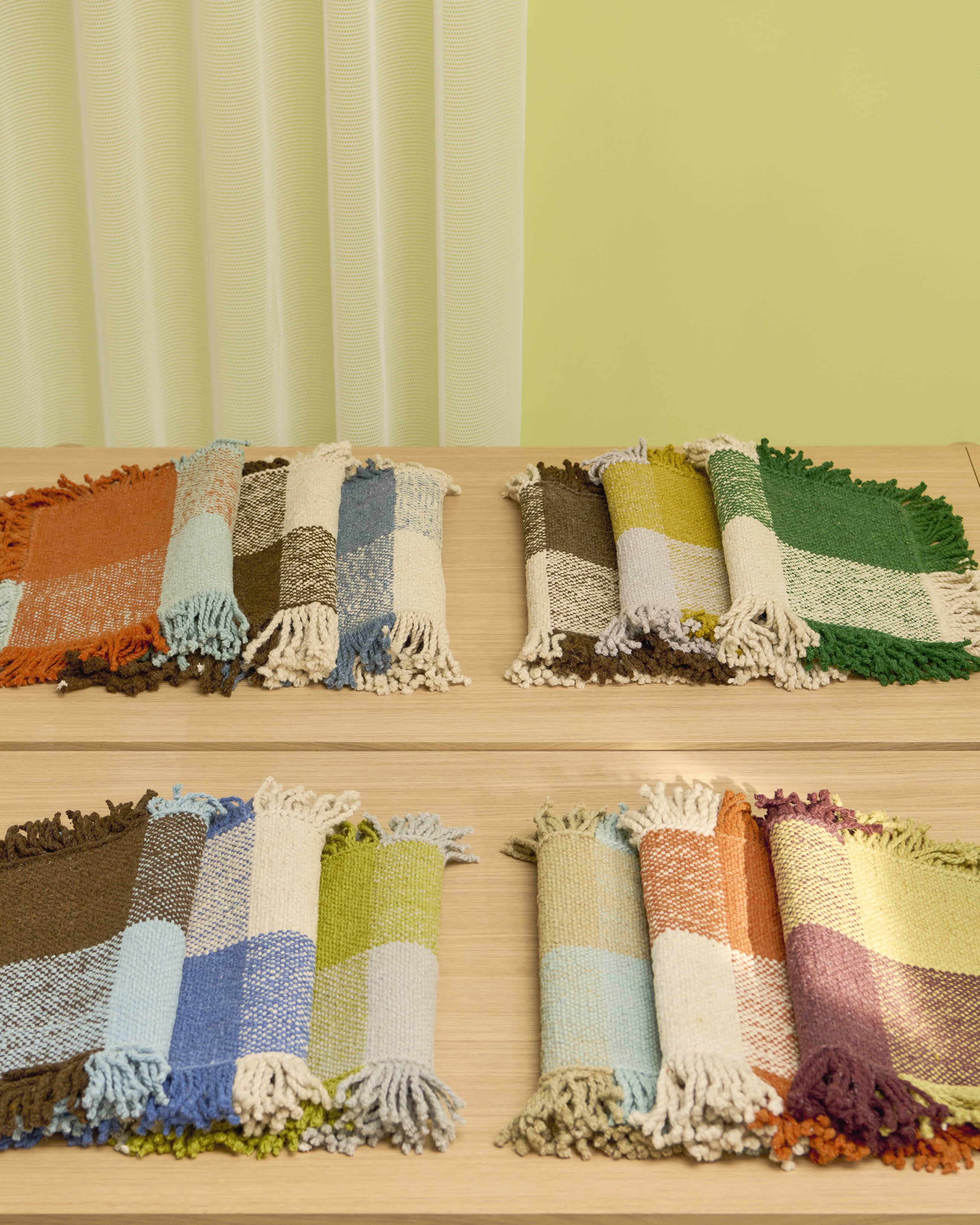 Four sets of handwoven Omni rug swatches  are neatly arranged on a light wooden table. Each swatch features bold color-blocked patterns with visible weaves and thick fringe edges in varied palettes, including earth tones, blues, greens, and oranges, against a soft lime green wall and sheer curtain backdrop.

