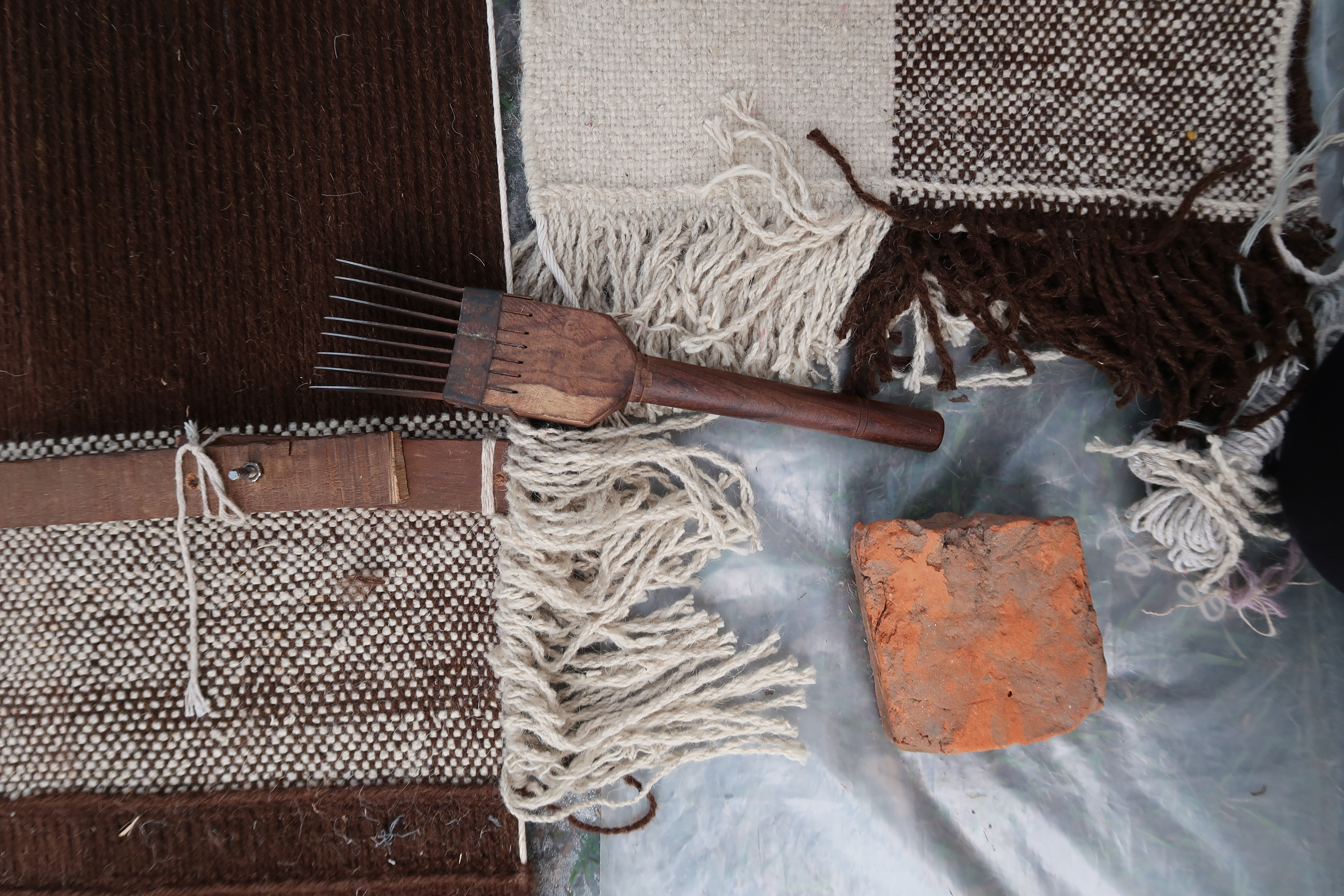 Detail of the Omni rug in the making, showing cream and brown sections with coarse fringe, a wooden weaving comb, a brick, and tools laid. The handcrafted nature of the rug is emphasized by the visible weft and rustic setting, reflecting traditional techniques.

