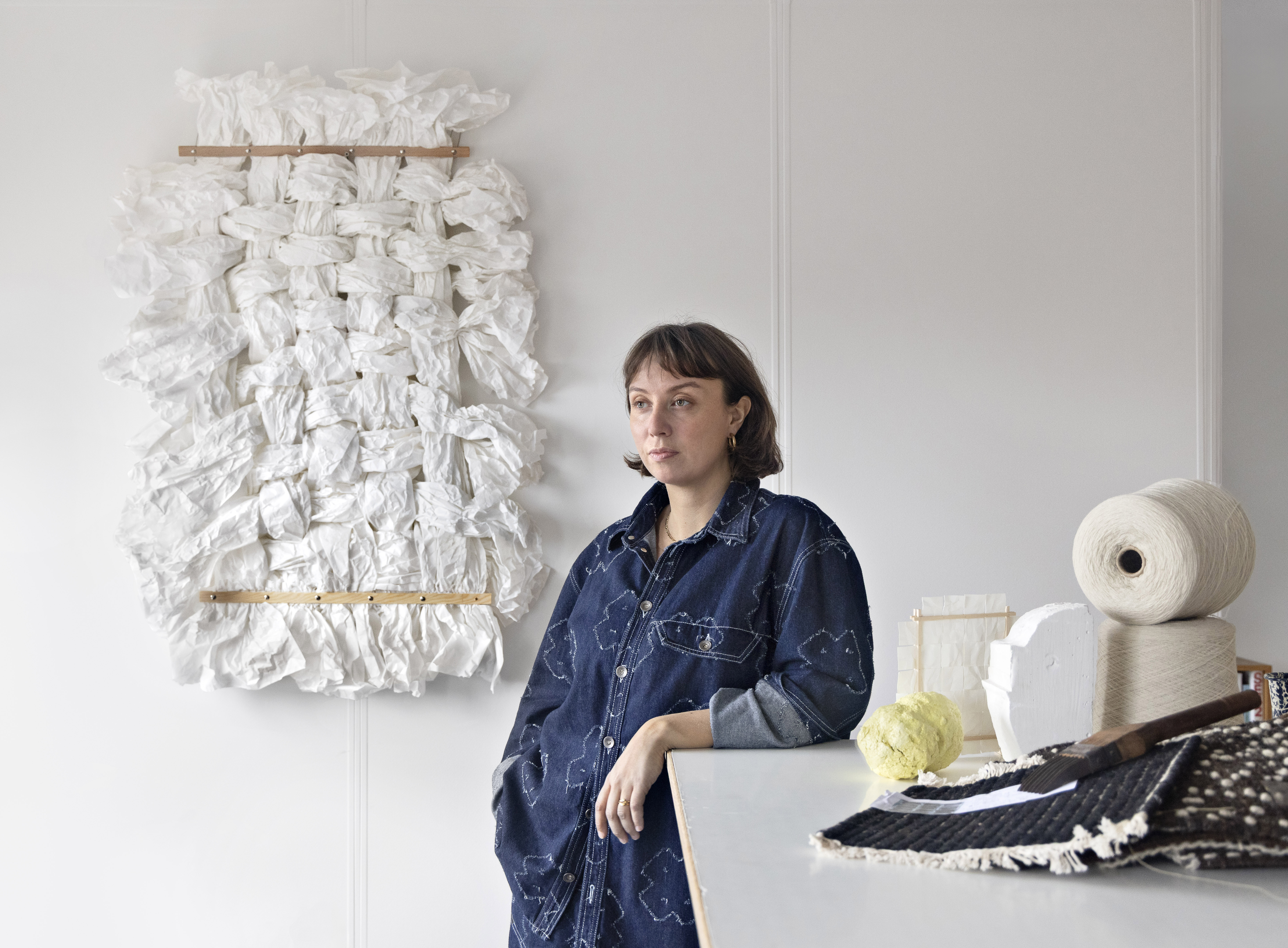 Danish designer Sia stands in her minimalist studio beside a worktable, dressed in a denim outfit with light embroidery. Behind her, a sculptural wall textile made of knotted and bunched white fabric hangs from wooden rods. On the table are crafting materials.