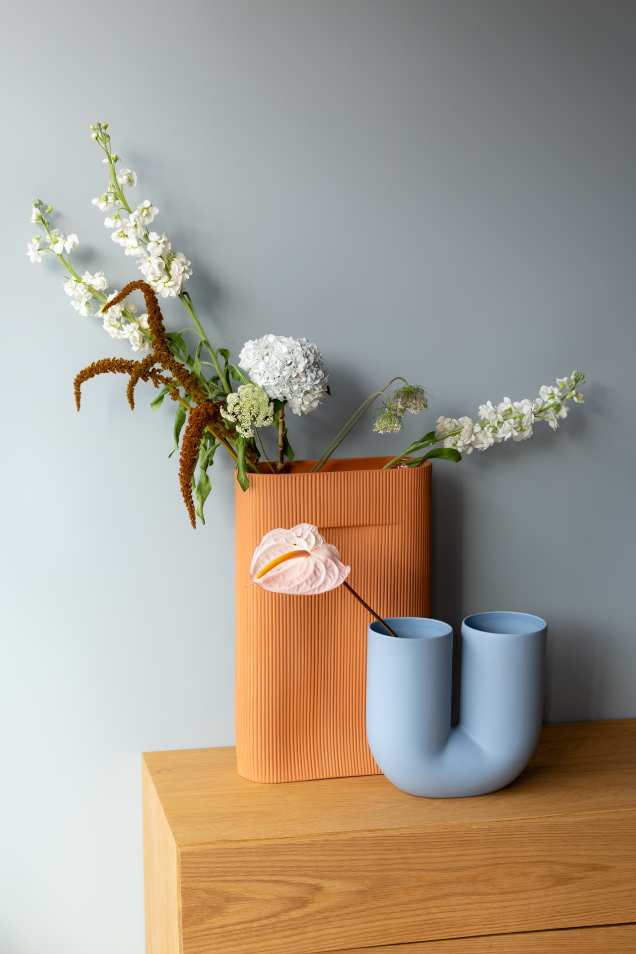Two sculptural vases, one in light blue and the other in terracotta, filled with colorful flowers, are placed on a wooden table.