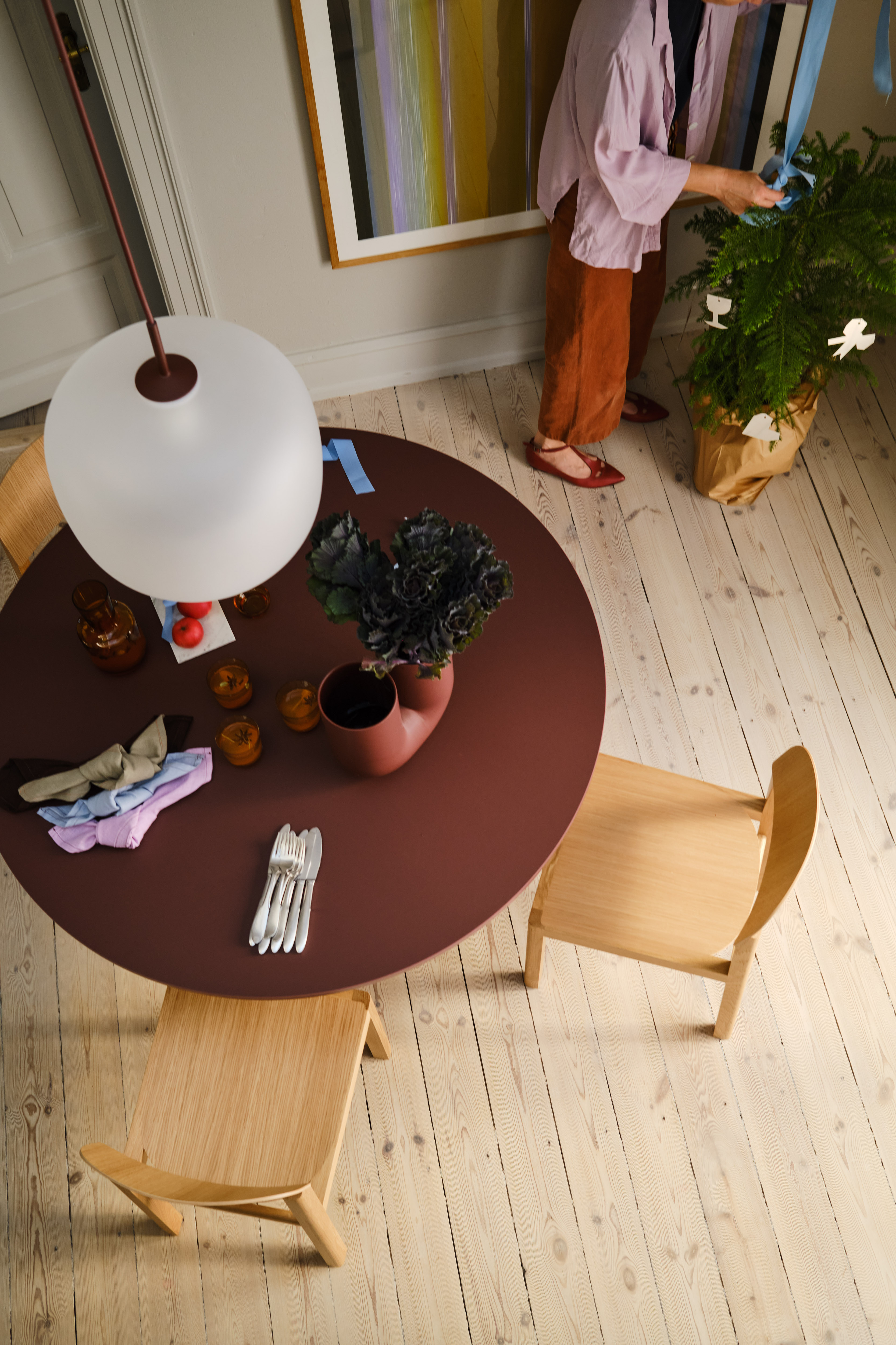 A woman in a lavender blouse arranges blue ribbons around a green potted plant. The table nearby features folded napkins, a deep red vase with kale, and a few glasses.